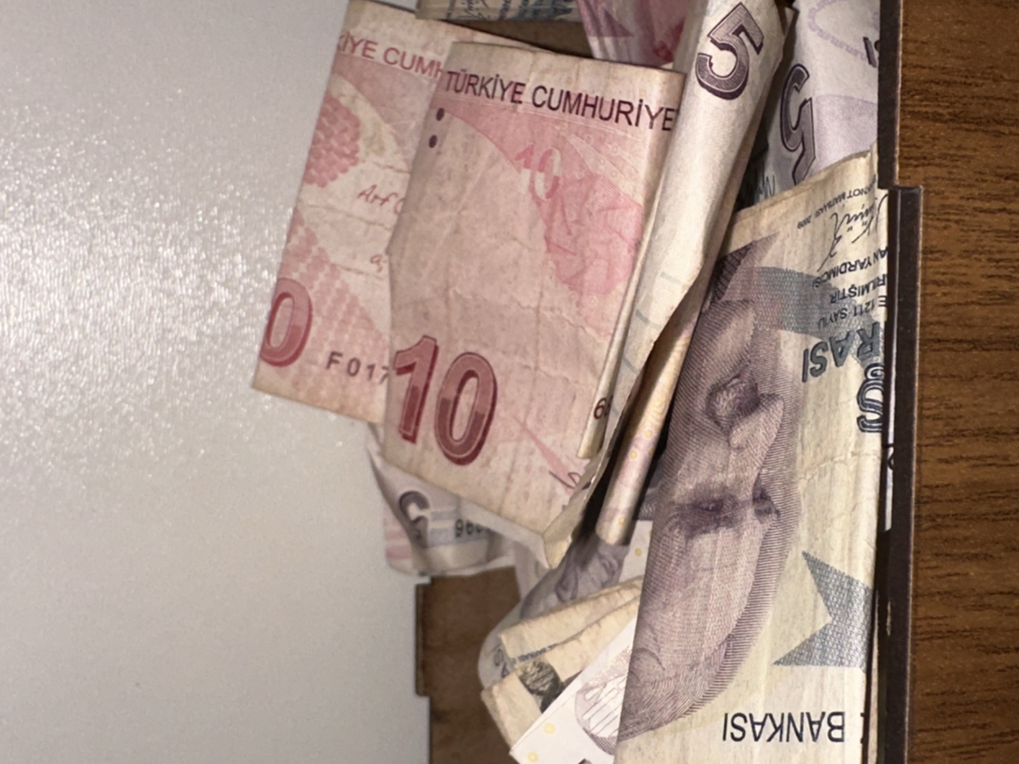 A pile of Turkish Lira banknotes is shown in a close-up, top-down view, resting on a wooden surface. The banknotes are varied in denomination, with visible numbers such as "10" and "5" printed on them. Some of the notes display text in Turkish, including "TÜRKİYE CUMHURİYETİ" and "BANKASI". The lighting suggests an indoor setting, likely a drawer or container, with the upper portion of the image showing a textured white wall or ceiling. The scene is devoid of any people, actions, or interactions. The image does not provide any visual cues to determine the specific location within Esenyurt, Turkey.