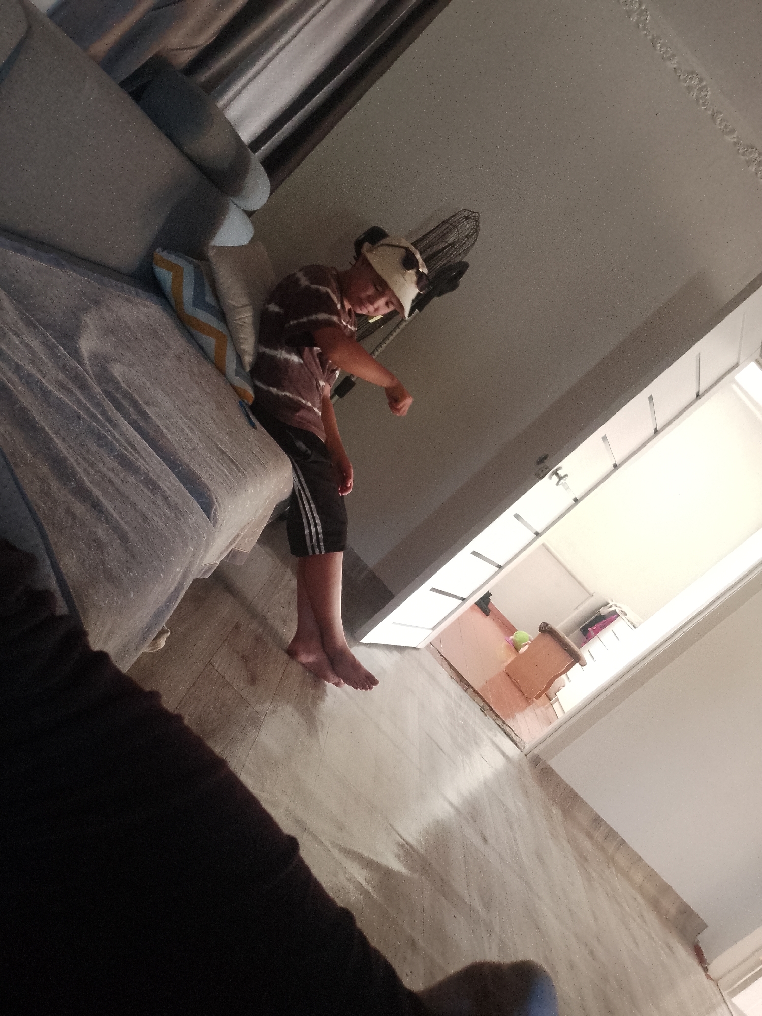 The image depicts a young boy sitting on a bed or couch in a room. He is wearing a striped t-shirt and dark shorts with white stripes. On his head, he has a novelty hat with animal ears. The boy is looking down and appears to be engaged in an activity, possibly playing a game, as his right hand is extended forward in a gesture. Behind him, a fan is mounted on the wall, and to his left, there are pillows on the couch. The floor is made of light-colored tiles. The lighting in the room is dim, with some natural light coming from a doorway in the background, which leads to another area with furniture. The overall scene suggests a casual indoor setting, likely a home. This image is not geolocated to any specific city or country, including Келдемұрат, Kazakhstan.