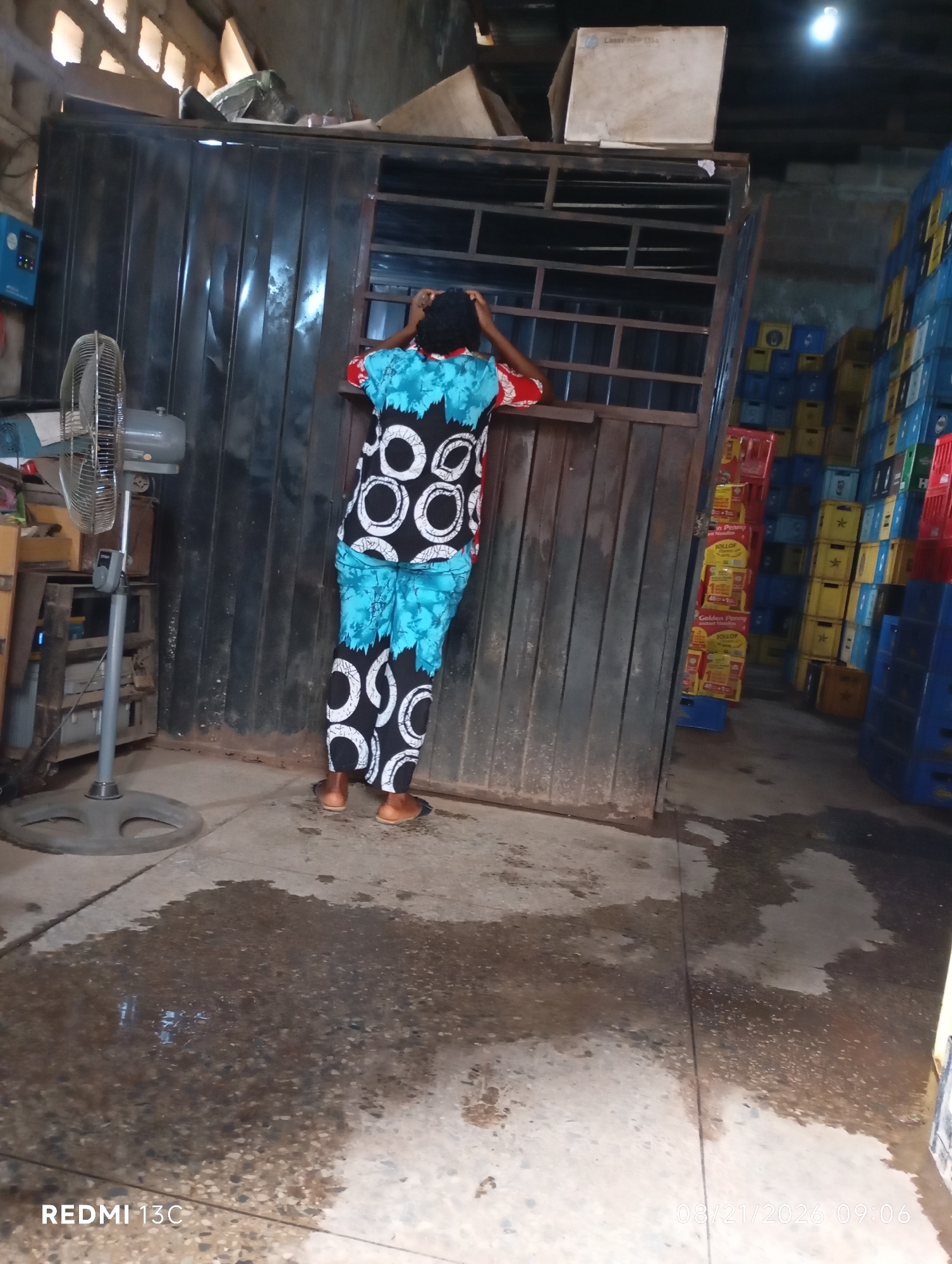 A person stands in a warehouse, facing away from the camera, with their hands on their head. They are wearing a blue and black patterned outfit. To the left, a gray oscillating fan stands on the floor. Behind the person, a metal structure with a window can be seen, and to the right, stacks of plastic crates filled with beverages are visible. The floor is wet and appears to be concrete. The lighting in the scene is dim, with a single lightbulb visible overhead. The overall impression is one of an industrial or storage setting.