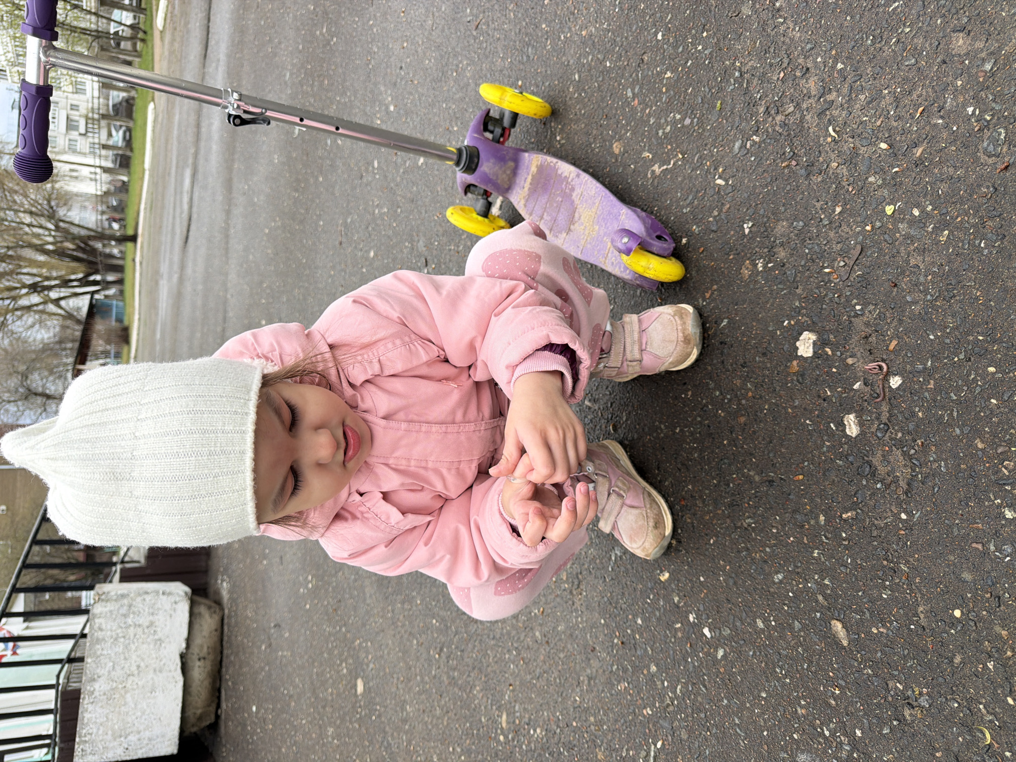 A young child, likely a toddler or preschooler, is seen squatting or kneeling on a damp asphalt surface in an outdoor setting. The image is captured from an elevated perspective, looking down at the child and their surroundings.
The child is dressed in warm clothing, including a thick, ribbed white knitted beanie hat, a pink padded jacket with an elasticated waist, and matching pink trousers adorned with small white polka dots. Their footwear consists of scuffed pink sneakers with white soles. The child's hands are clasped together in front of them, appearing to be focused on their fingers or a small object. Their eyes are cast downwards, and their mouth is slightly open, conveying a calm or contemplative expression.
Adjacent to the child is a kick scooter. The scooter has a purple deck and bright yellow wheels, with a silver metallic handlebar that features purple grips. The scooter shows signs of wear and dirt, indicating it has been well-used.
The ground is a dark, coarse asphalt pavement, appearing damp or recently wet, with scattered pebbles and small pieces of debris. In the blurred background, partially visible elements include bare trees and distant, light-colored multi-story buildings, characteristic of an urban or suburban landscape in a place like Chaikovsky, Russia. The overall lighting suggests an overcast or dull day, possibly in early spring or late autumn given the bare trees.
There is no visible text in the image.