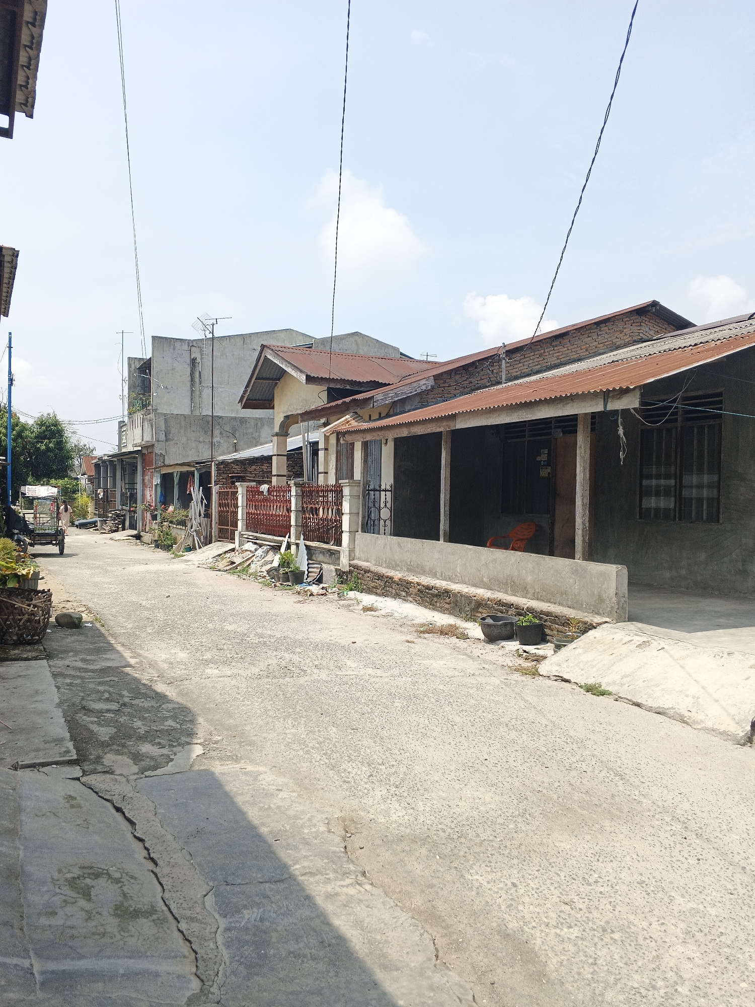 This image depicts a street scene in the city of Tanjung Morawa, Indonesia. The street is paved with concrete and shows signs of wear, with visible cracks and uneven surfaces. On either side of the street are residential buildings with various architectural styles, predominantly featuring simple construction with corrugated metal roofs and concrete walls.
On the left side of the street, a person is pushing a cart laden with goods, possibly a vendor. The cart is a two-wheeled vehicle with a covered top. A large woven basket is placed on the ground near the cart, containing plants or other items. Further down the street, more houses and some trees are visible.
The right side of the street features a row of houses with gates and fences. One house has a porch area with an orange chair visible inside. There are potted plants and some debris scattered along the property lines. Electrical wires are strung across the scene, supported by poles. The sky is bright blue with some scattered clouds, indicating a daytime scene. The overall impression is that of a typical residential street in a developing urban or suburban area.
