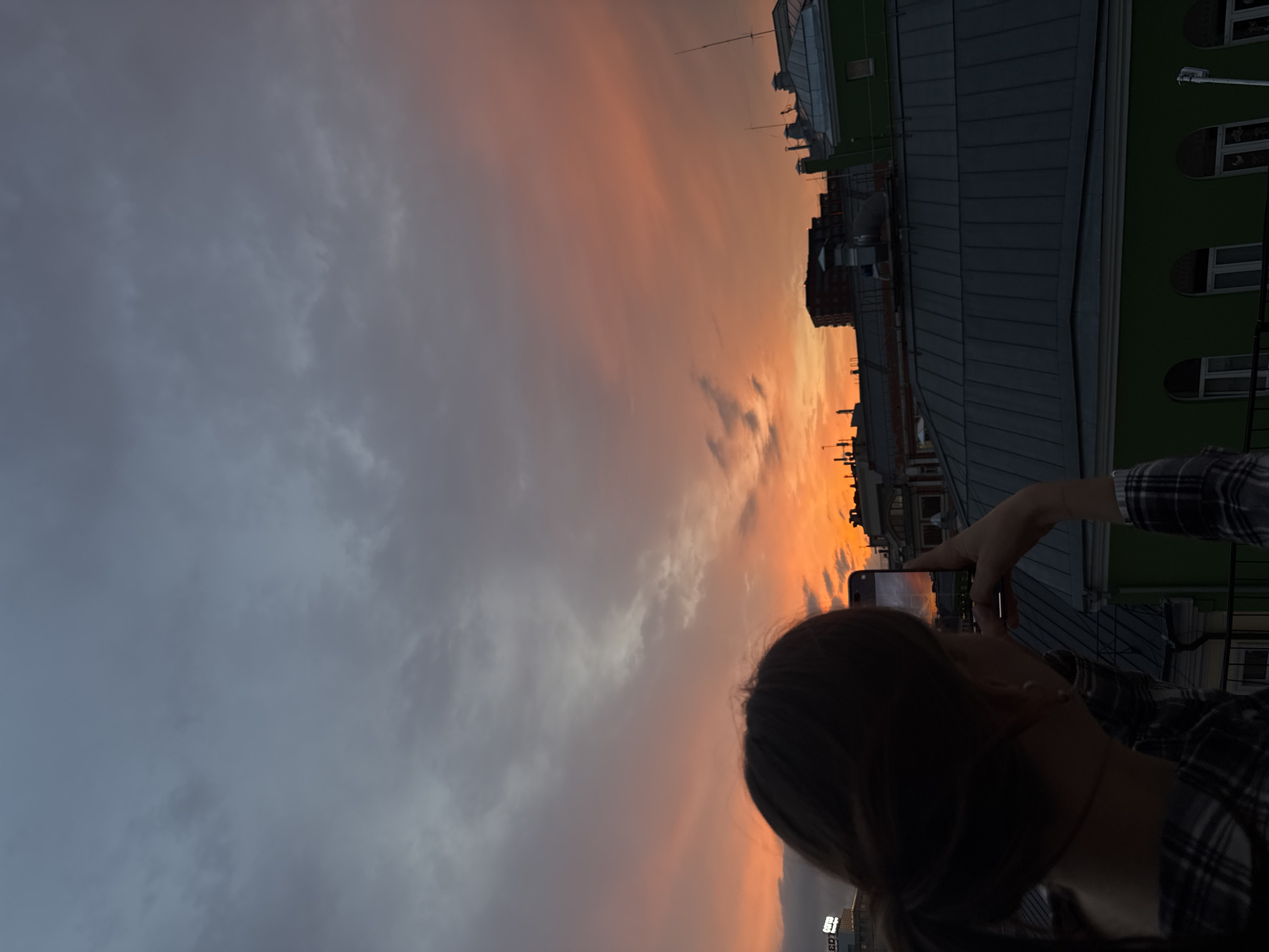 A woman is holding a smartphone to photograph a sunset sky. The sky is a mix of dark grey clouds and orange and pink hues. In the distance are buildings, likely in Moscow, Russia. The woman is wearing a plaid shirt.