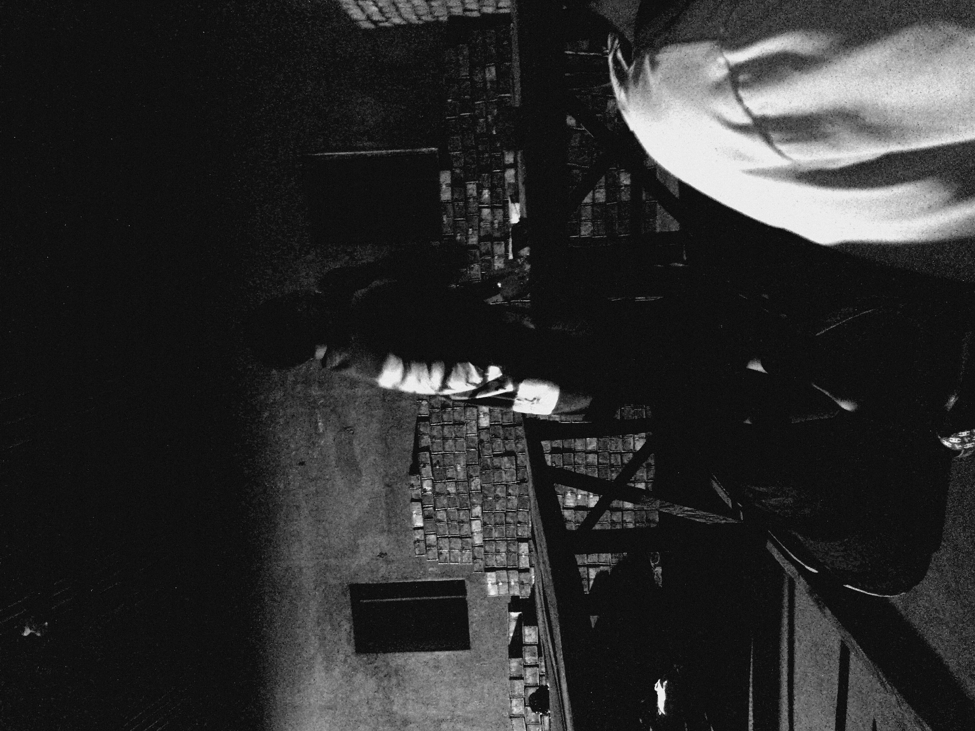 The image is a high-angle, black-and-white photograph depicting a city street at night. The environment is mostly dark with limited illumination, creating high contrast. The street appears to be constructed with small, square stones. There is a dark, rectangular opening in the street. A figure wearing a white garment is visible on the right side of the frame. Wooden structures are present on the street, including a cross-shaped frame.