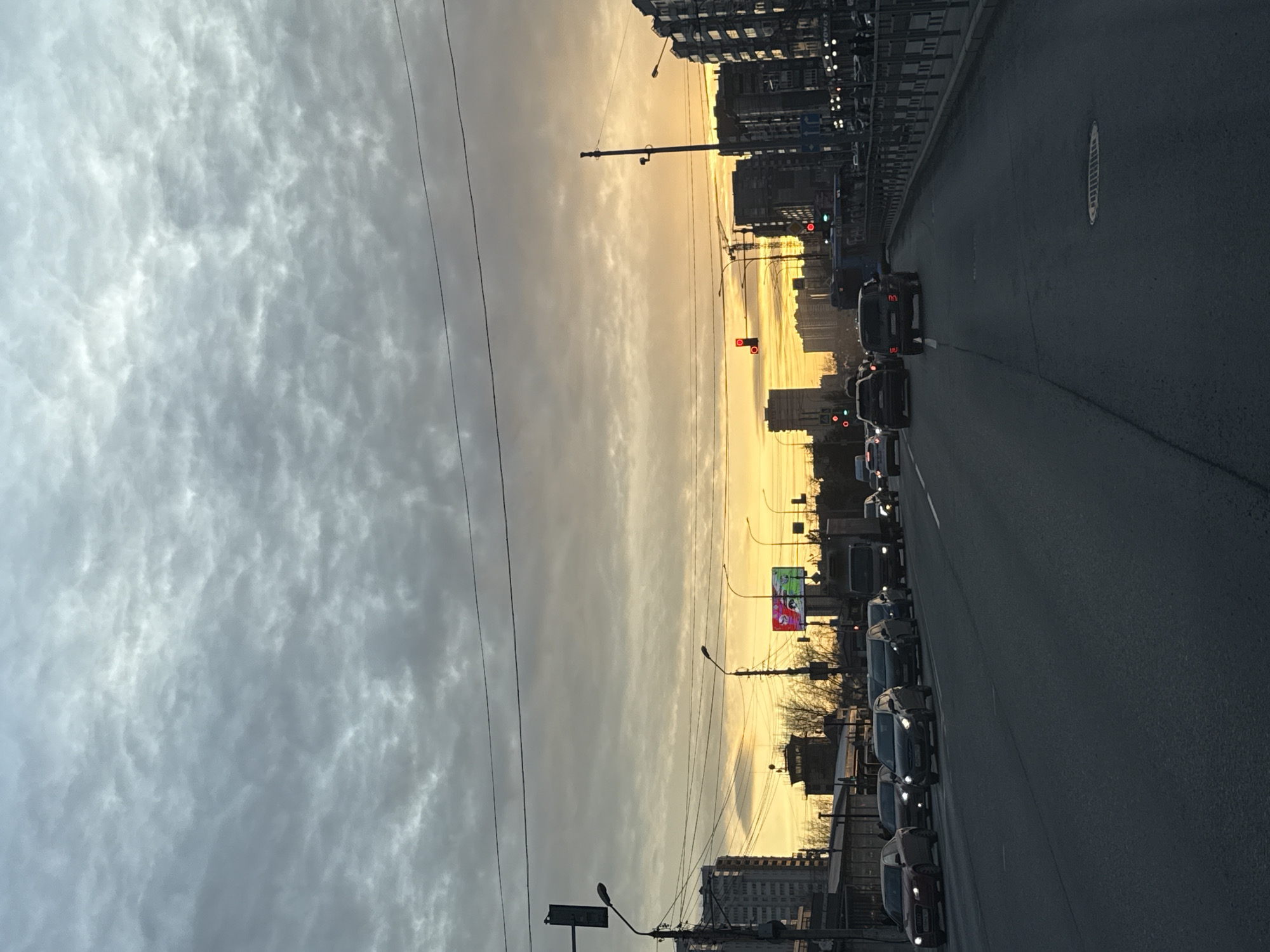 The image depicts a city street at dusk with a line of cars waiting in traffic. The sky is a mix of grey clouds on one side and an orange and yellow glow from the setting sun on the other side. Buildings are visible on both sides of the street. Power lines run across the sky. Red traffic lights are illuminated in the distance. The photograph was taken in Krasnoyarsk, Russia.