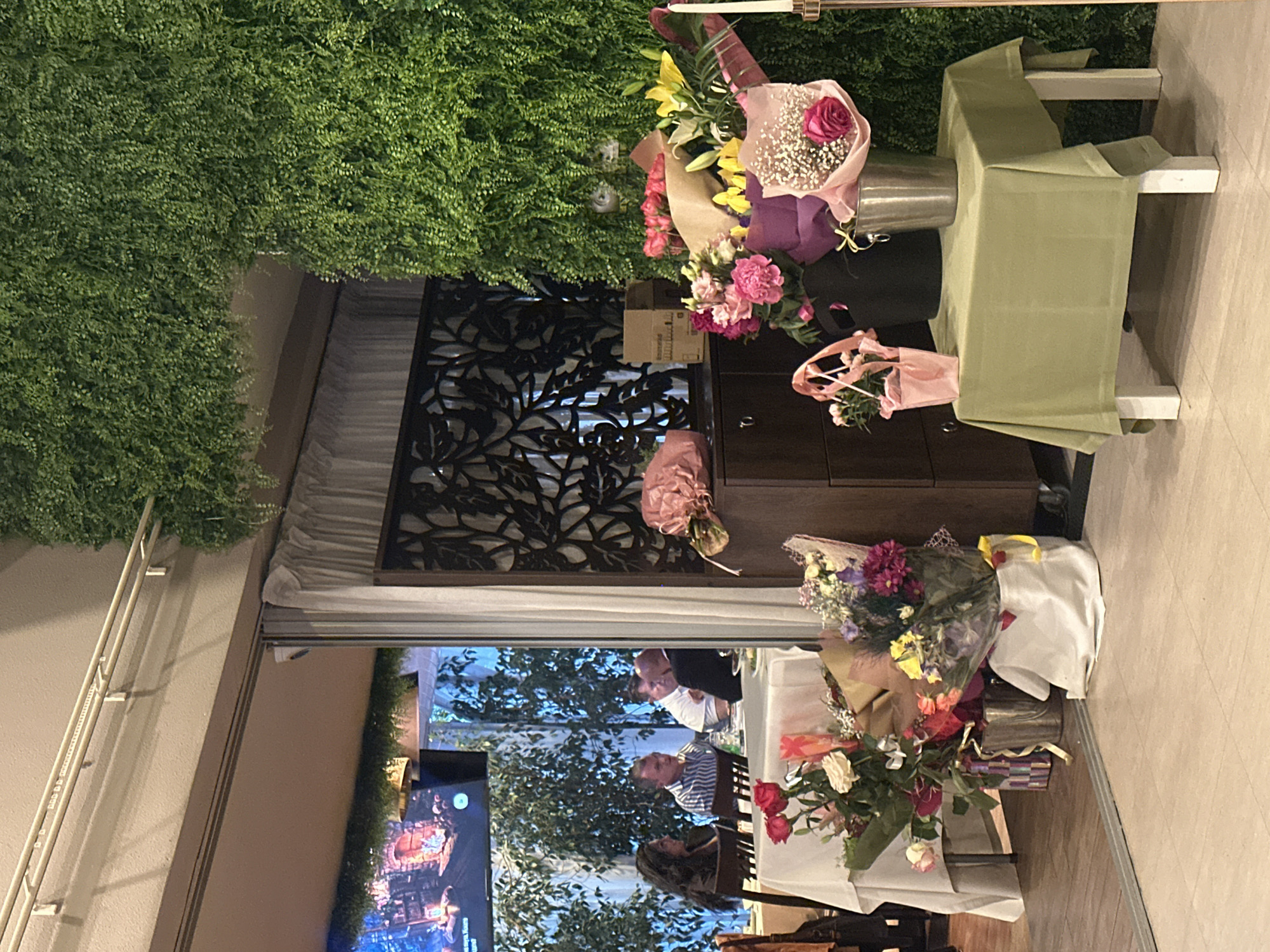The image shows an interior scene, likely a restaurant or event space. There are tables set for a gathering with people visible through a doorway. Several bouquets of flowers are prominently displayed, some on a table covered with a light green tablecloth. There is a dark wooden cabinet behind the flowers. A television screen displays an image with bright colors. The room is decorated with greenery on the walls. The precise location within Sofia, Bulgaria cannot be determined from the image. The people visible appear to be casually dressed and seated at tables. The overall impression is one of a celebratory event or social gathering.