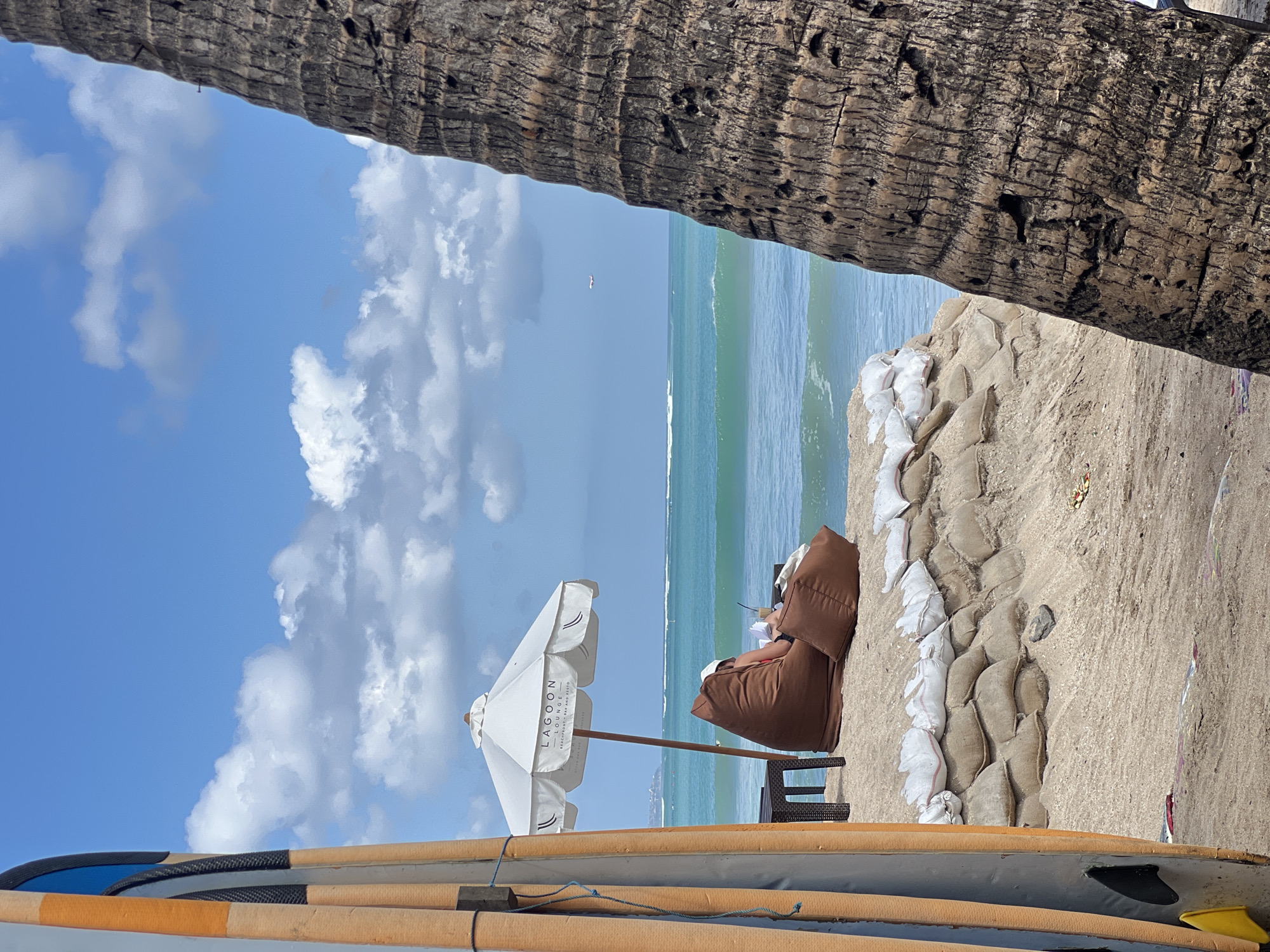 The image depicts a scene on a beach in Kuta, Indonesia. In the foreground, on the left, are two surfboards standing upright, leaning against each other. To the right of the surfboards, a person is seated on a large brown beanbag chair under a white umbrella. The umbrella has the text "LAGOON LOUNGE BEACHFRONT BAR & RESTO" printed on it. The person is holding a book and a drink with a straw. A small brown table with a dark surface is positioned to the left of the beanbag chair. Behind the person and the beanbag, a wall constructed of sandbags is visible. The beach is sandy, and the ocean with gentle waves stretches out to the horizon. In the distance, an airplane is visible in the sky. A portion of a large palm tree trunk is prominent on the right side of the image, obscuring part of the background. The sky is blue with scattered white clouds.