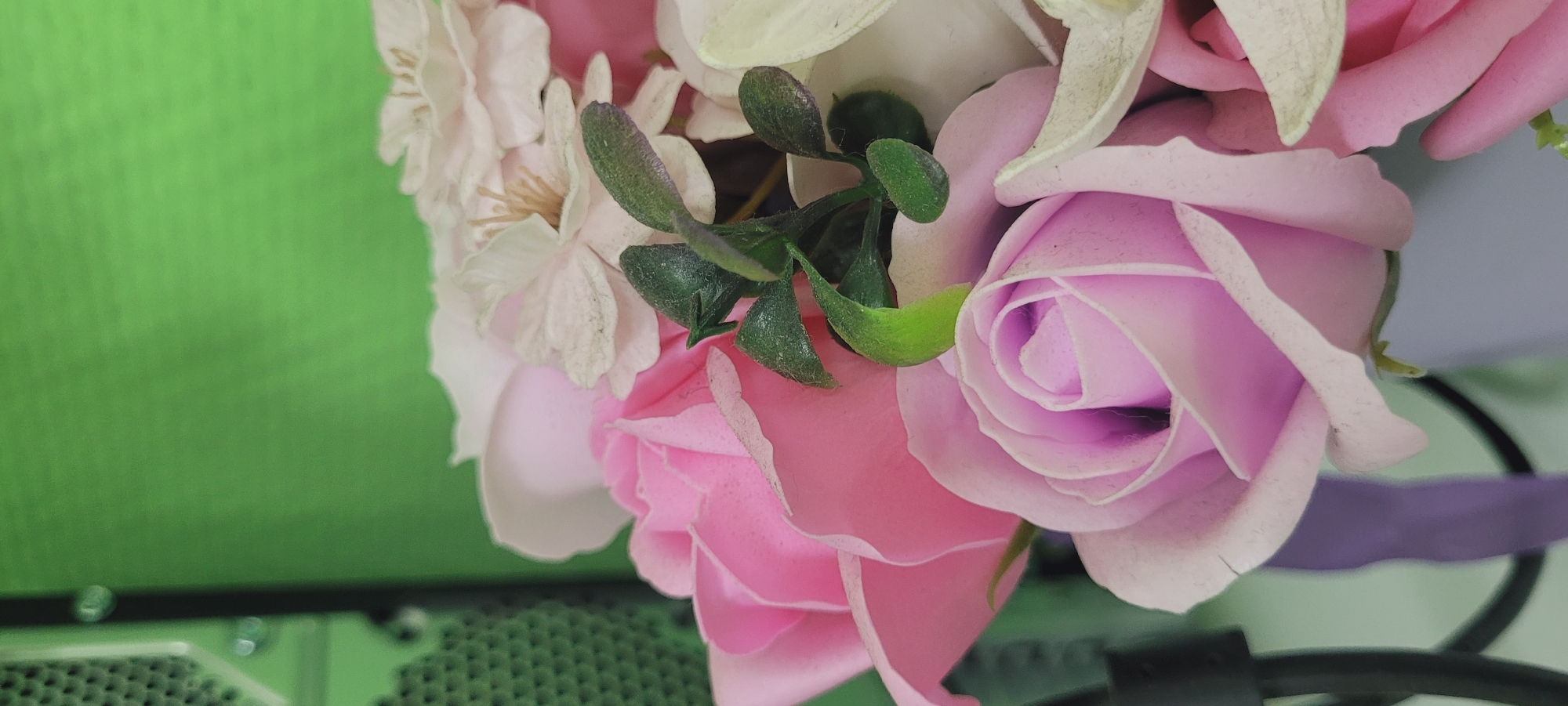 The image depicts a close-up of a bouquet of artificial flowers, predominantly in shades of pink and white, with some green foliage. The flowers include roses and other smaller, delicate blossoms. The arrangement is set against a blurred background of green and a partial view of what appears to be a black metal object.