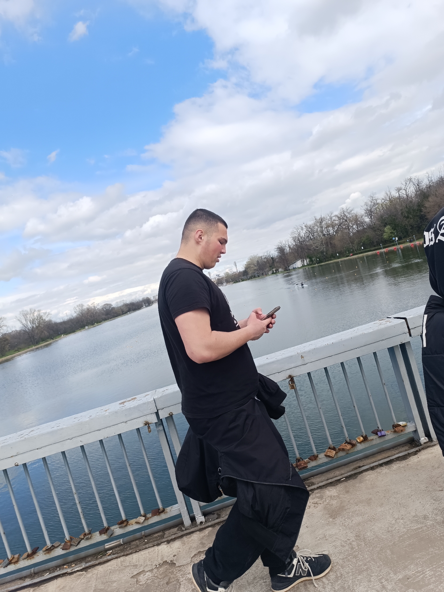 A medium shot captures a man, likely in his twenties, standing on a bridge and looking at a mobile phone. He is wearing a black t-shirt, black trousers, and black and white trainers. A black jacket is draped around his waist. He is positioned to the left of the frame, with a portion of another individual visible on the right, wearing a black hooded top with white writing, and black trousers.
The bridge has a gray railing adorned with numerous padlocks. Behind the bridge is a calm lake with a few rowboats visible in the distance. The far bank of the lake is lined with trees, partially bare, indicating either early spring or late autumn.
The sky is partly cloudy with patches of blue.
This is likely the rowing canal in Plovdiv, Bulgaria.