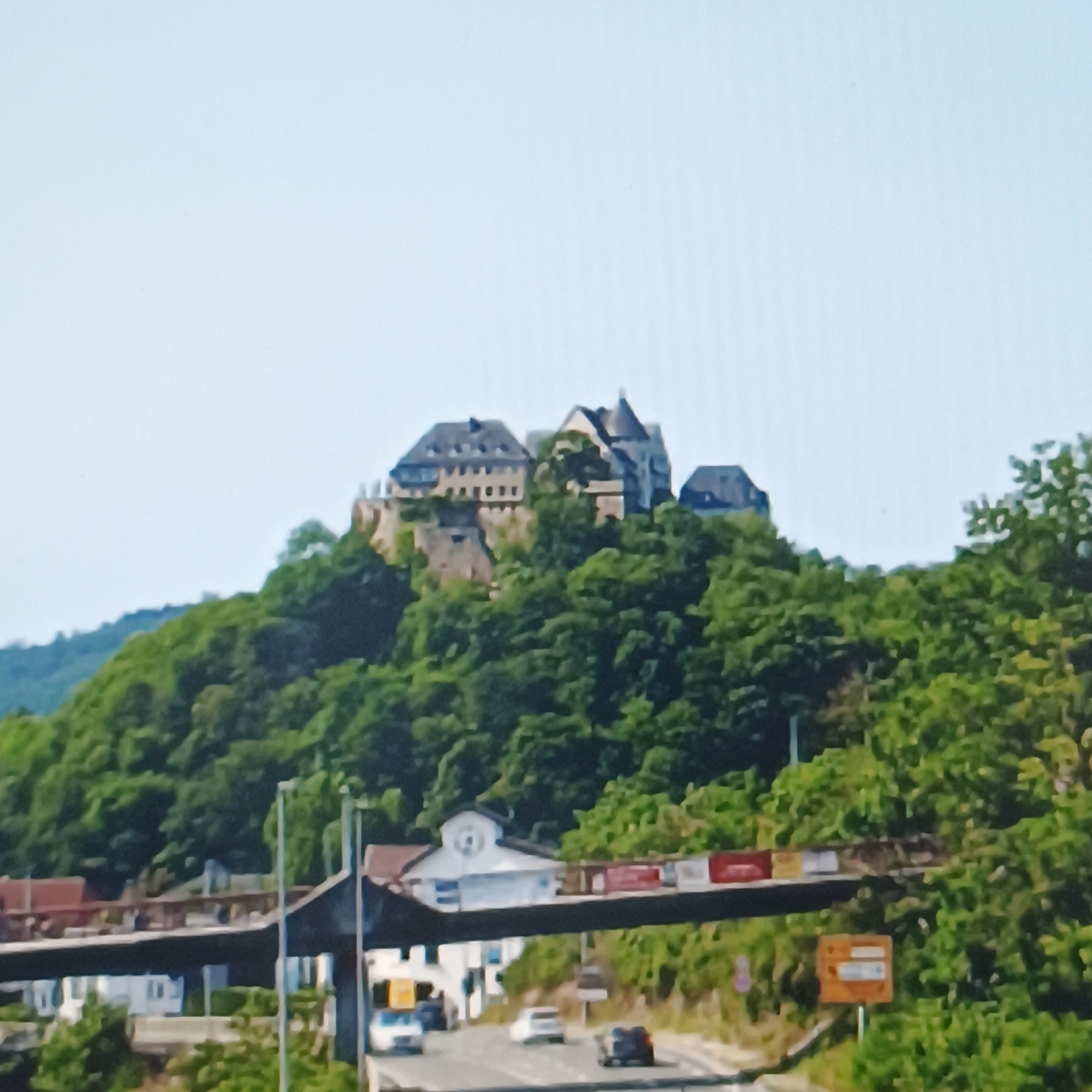 The image captures a scenic view of Rüsselsheim am Main, Germany, featuring a prominent castle situated atop a lush, tree-covered hill. In the foreground, a multi-lane road passes beneath a bridge-like structure adorned with signage. Several vehicles are visible on the road, suggesting active traffic. The castle, with its distinctive architecture including multiple rooflines and stone walls, is the focal point of the upper portion of the image. The surrounding vegetation is dense and green, indicating a natural, possibly rural or park-like setting within the city. The sky is a clear, pale blue.