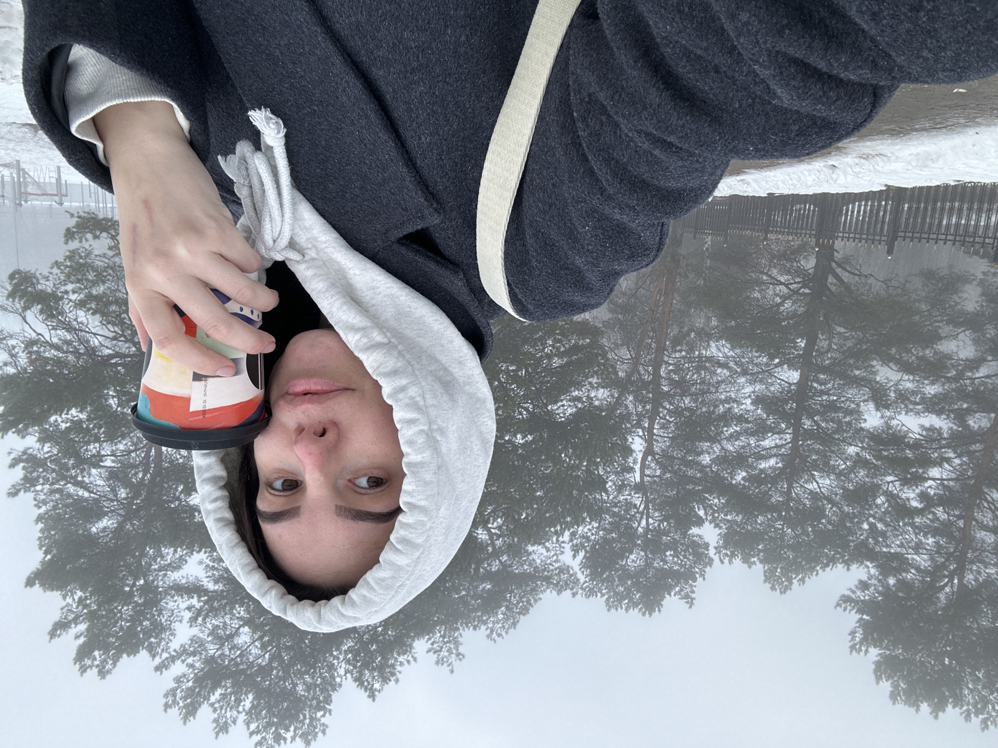 The image shows a person in Chaikovsky, Russia, holding a disposable coffee cup. The person is wearing a gray hoodie, a dark gray coat, and a light-colored shirt underneath. A light-colored strap is visible on their shoulder. The background includes snow-covered ground, a fence, and trees shrouded in fog.