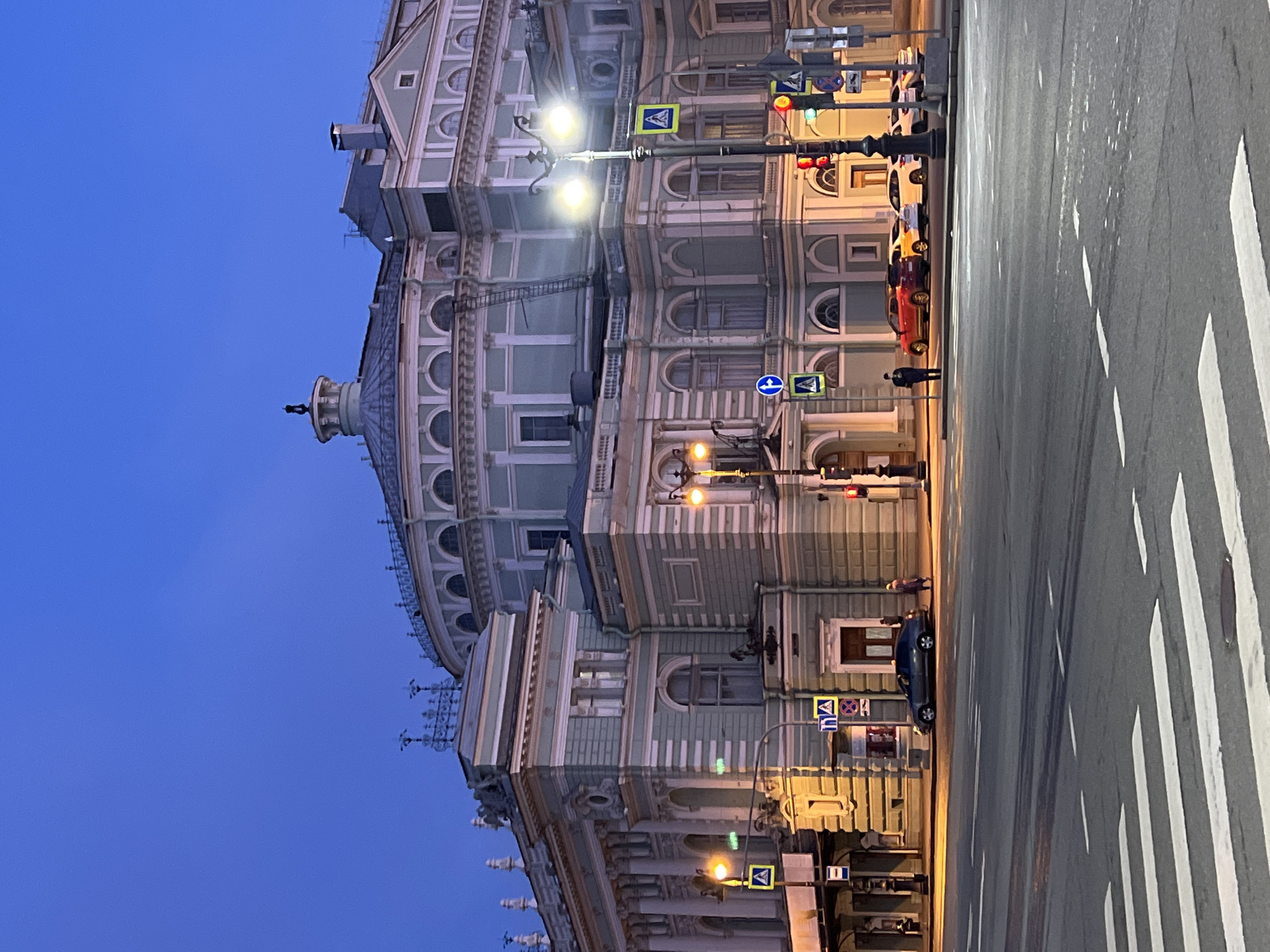 The image depicts a building in Saint Petersburg, Russia. It is a grand, multi-story structure with a pale green facade, featuring arched windows and classical architectural details. A dome sits atop the building. Streetlights illuminate the building, casting a warm glow on its surface. Traffic lights are visible along the roadside. The street is wide and includes a crosswalk. Several cars are parked along the side of the road. A person is walking on the sidewalk. The sky is a deep blue, suggesting it is dusk or dawn. The overall atmosphere is urban and stately.