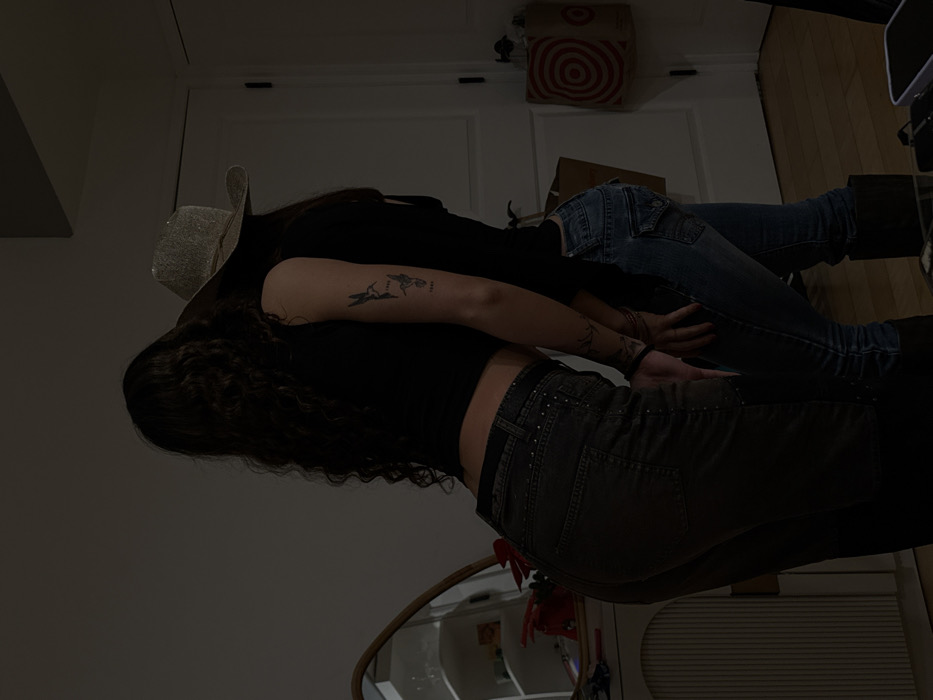 Two women lie on the floor. One, wearing a cowboy hat, leans on the other. They both wear black tops. The woman in the hat has a tattoo on her arm. The other woman has dark curly hair. They both wear jeans. A Target bag is on the wall in the background. A mirror and radiator are also visible.