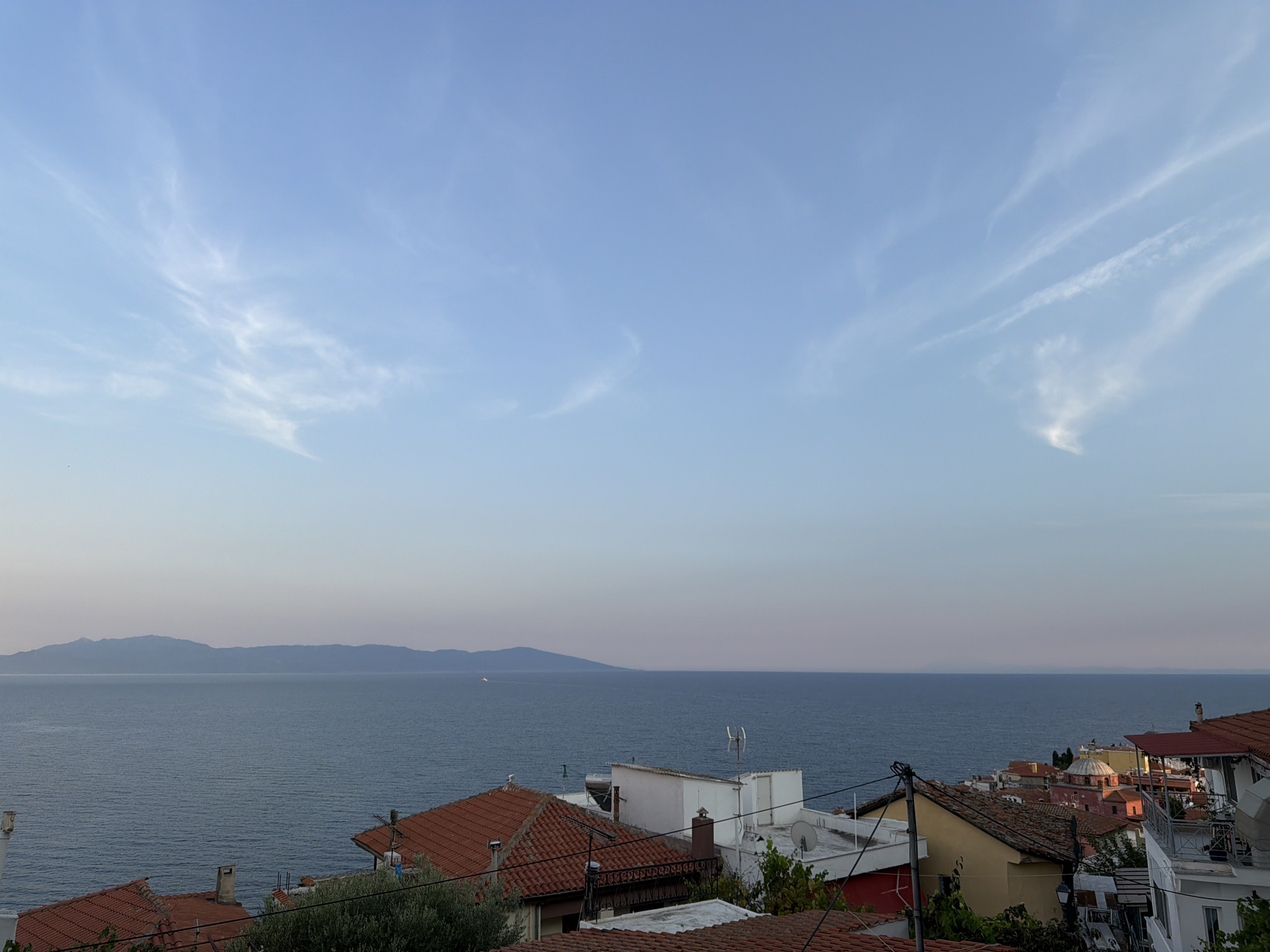 The image captures a panoramic view of the Aegean Sea from a residential area in Kavala, Greece. The sky is a clear, pale blue with wispy white clouds stretching across it. In the distance, a long, hazy mountain range forms the horizon. The foreground is dominated by the rooftops of houses, characterized by their terracotta tiles. These buildings appear to be situated on a hillside, descending towards the sea. Some buildings have white facades, while others are painted in warm yellow and orange hues. Various architectural elements are visible, including chimneys, satellite dishes, and balconies. The sea itself is a deep blue, with gentle ripples indicating a calm day. A small, distant boat is visible on the water. The overall impression is one of a serene coastal town at dusk or dawn.