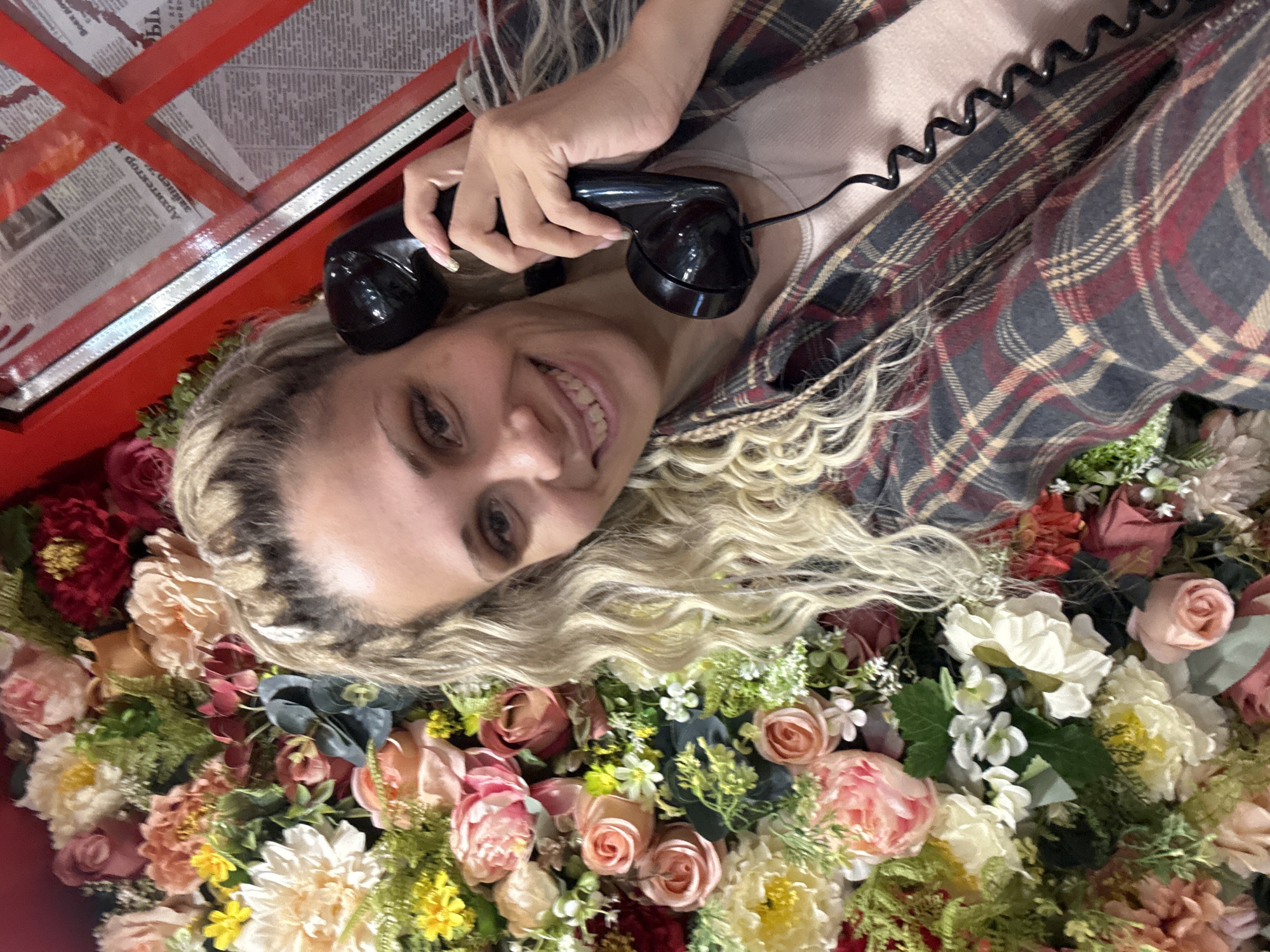 A woman is lying on a bed of flowers while holding an old-fashioned black telephone receiver to her ear inside a red telephone booth. The floral arrangement includes roses, peonies, and other assorted blooms in shades of pink, white, yellow, and red, interspersed with greenery. The woman has blonde hair styled in waves and is wearing a plaid shirt over a light-colored top. The telephone booth contains newspaper clippings on the back wall. There is no immediate indication that this is located in Шебунино, Russia.