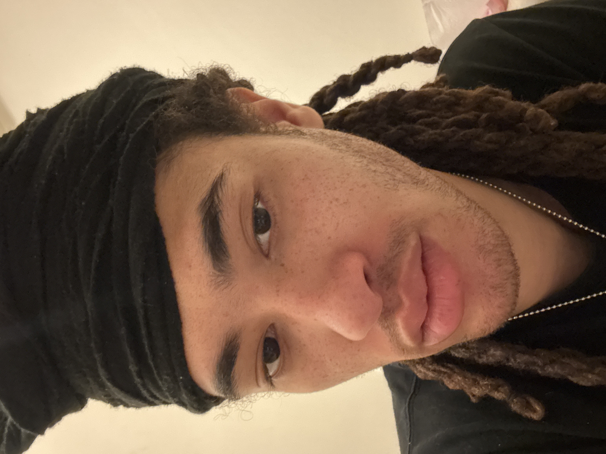 A close-up portrait features an individual with light brown skin, prominent freckles, dark eyes, and full lips, looking directly at the camera. The person has dark dreadlocked hair, with some strands visible around the face and shoulders, and wears a black fabric head wrap that covers the top of their head. Faint stubble is present on the chin and jawline. A dark garment and a silver ball-chain necklace are visible. The background is a plain, light-colored wall, indicating an indoor environment. No visual details within the image identify the location as Paris, France, or any specific city.