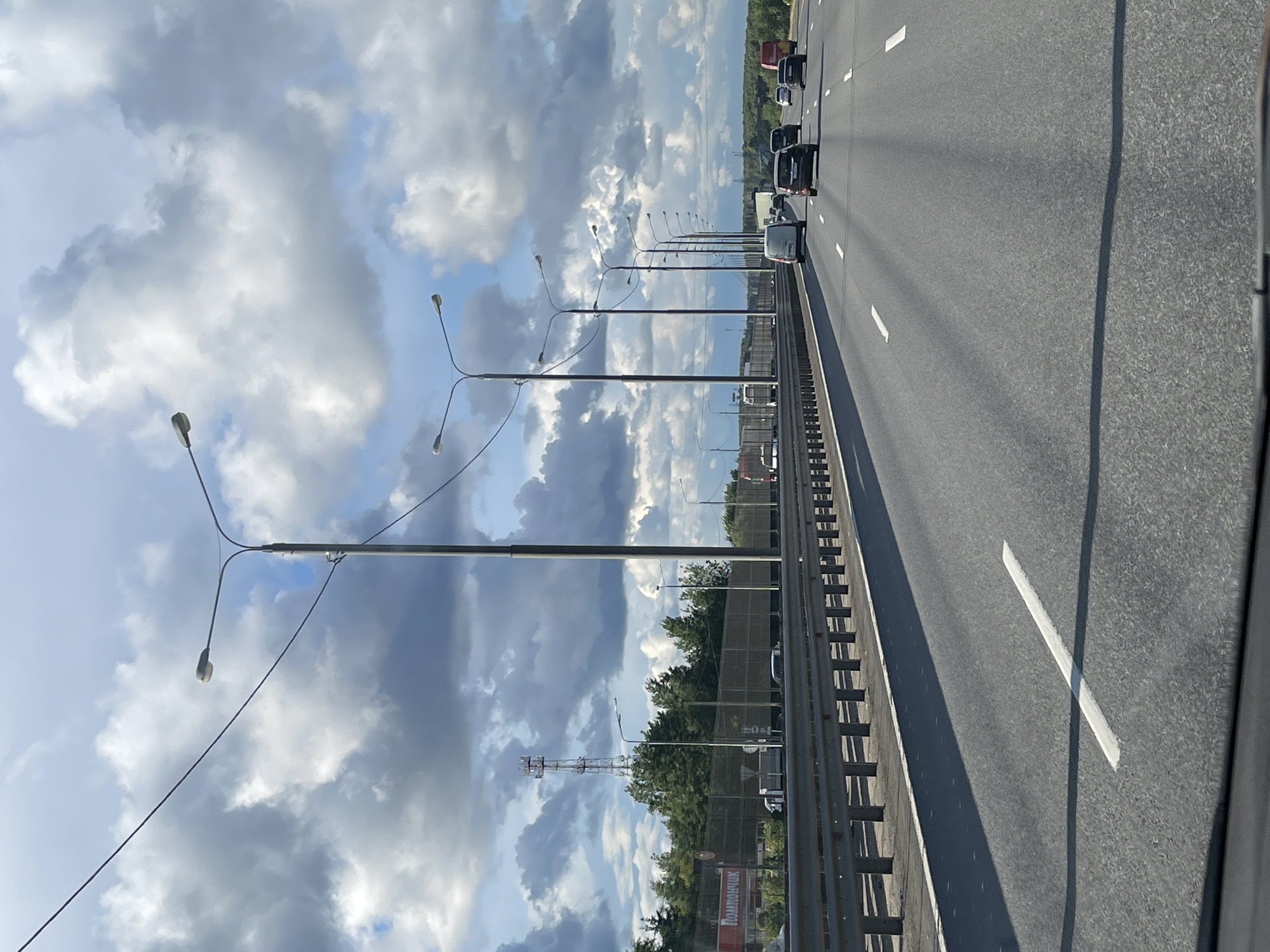 The image shows a highway in what appears to be Berezhki, Russia. The highway is lined with streetlights. On the left side of the road is a guardrail. Traffic is visible on the highway. In the background, trees are visible and a cell tower can be seen. The sky is filled with cumulus clouds.