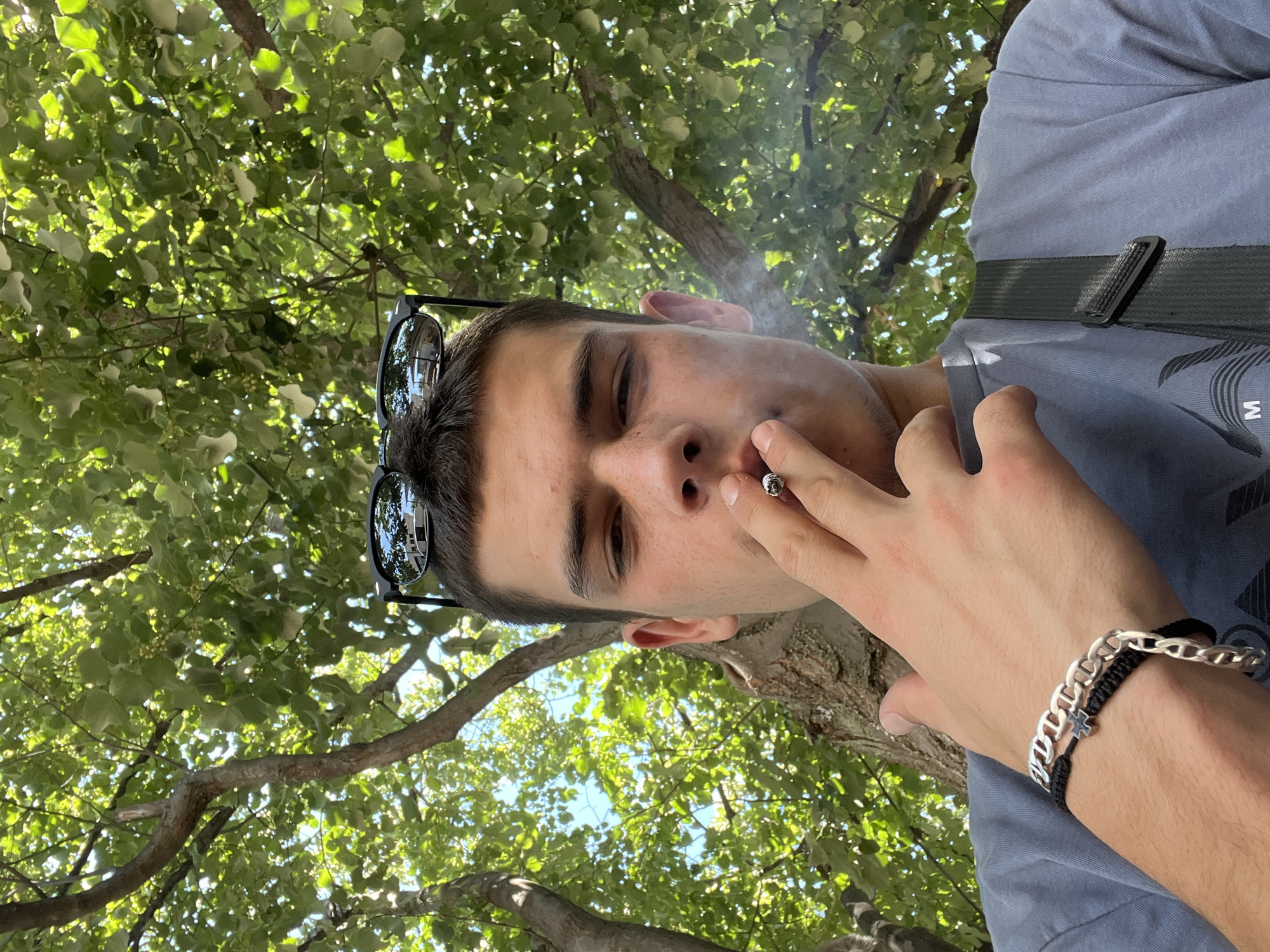 A young man, appearing to be in his late teens or early twenties, is captured in a close-up, low-angle selfie. He has short brown hair and is looking directly at the camera. Sunglasses are perched on top of his head. He is holding a lit cigarette to his lips, exhaling a plume of smoke. A silver chain bracelet adorns his left wrist, layered over a black braided cord bracelet. He is wearing a blue-grey t-shirt. A black strap of a bag is visible across his shoulder. The background is dominated by lush green foliage and tree branches, suggesting an outdoor setting, possibly in a park or a shaded area within the city of Sopot, Bulgaria. The sunlight filters through the leaves, creating a dappled effect.