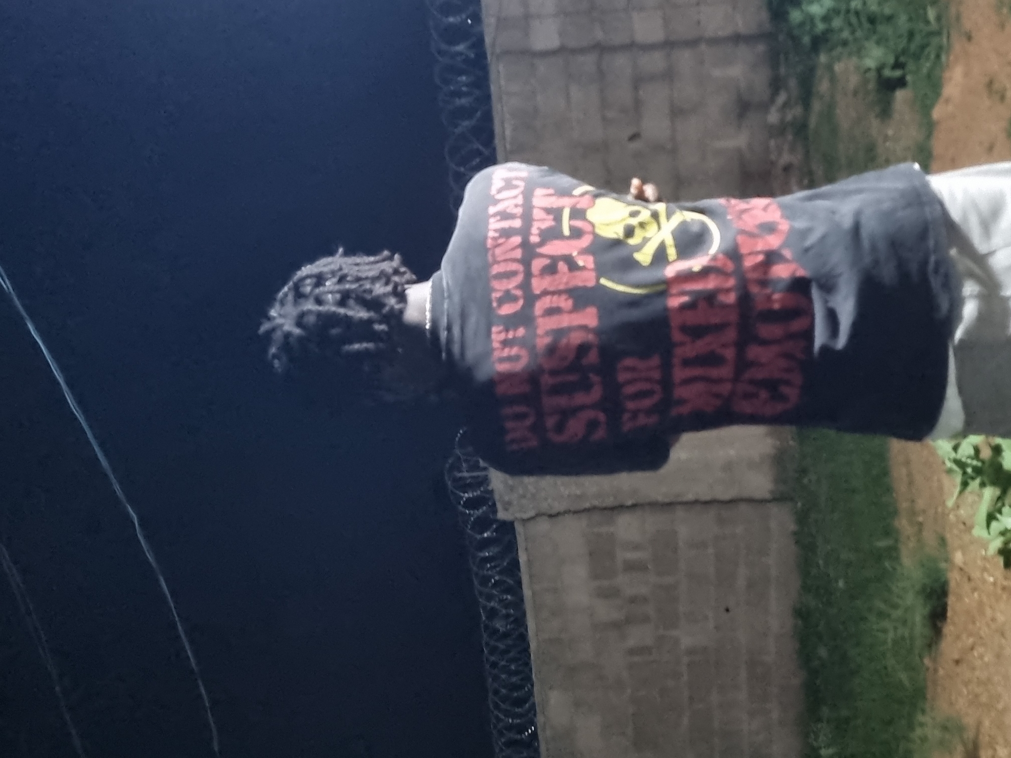 At night in Bar Jirgi Summa, Nigeria, an individual with braided hair wears a dark-colored T-shirt with a yellow skull and crossbones graphic above red text that reads "DO NOT CONTACT SUSPECT FOR MIXING EMOJI." The individual stands next to a low brick wall topped with coiled razor wire. The scene includes dark sky and foliage in the background.