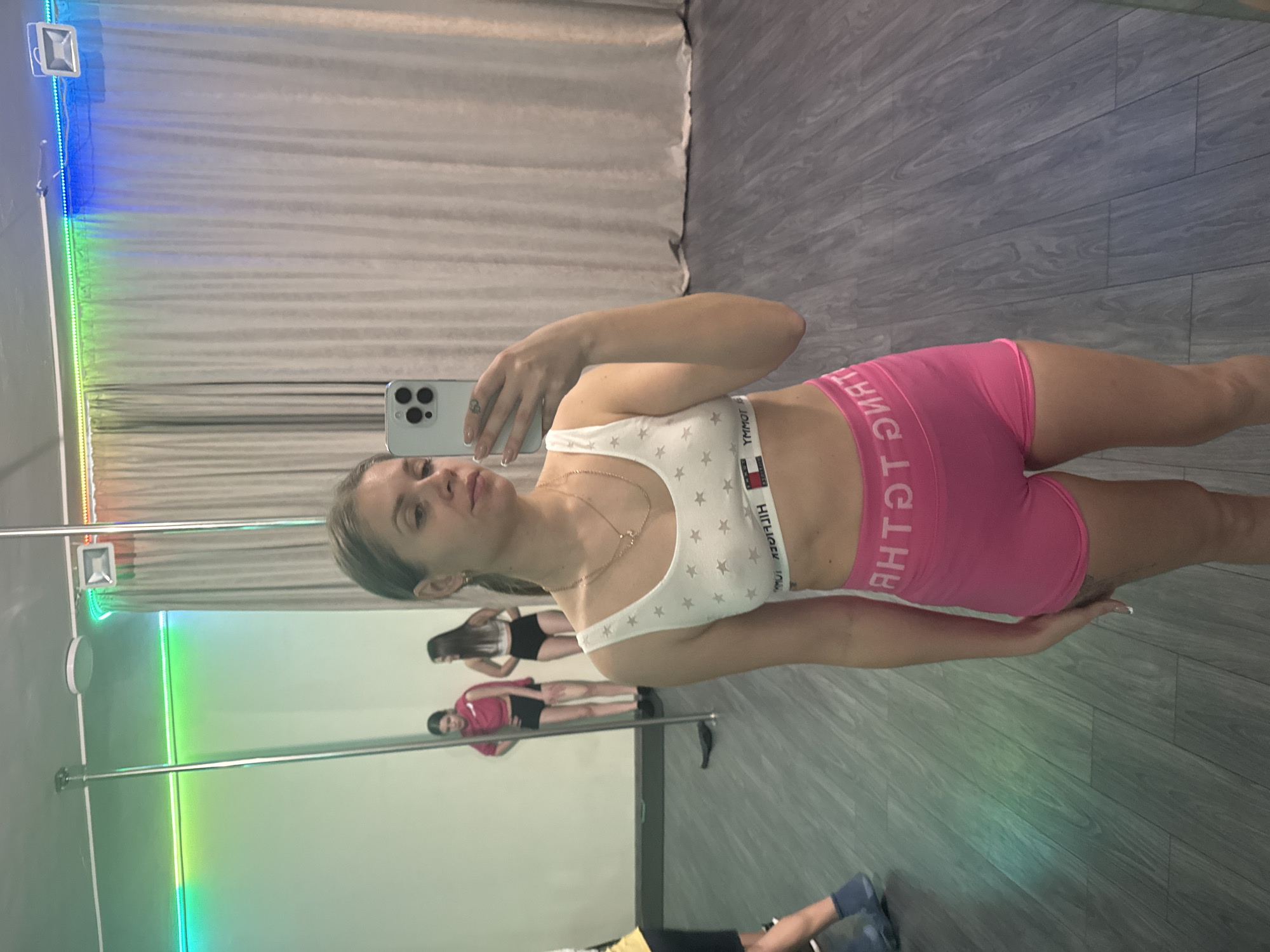 A woman is taking a selfie in a dance studio. She is wearing a white sports bra with star patterns, pink shorts, and a gold necklace. The studio has mirrored walls, with at least two other individuals visible in the reflection engaging in what appears to be pole dancing or exercise activities. The environment includes poles set up for dancing, and the lighting is a combination of natural and artificial, with a rainbow effect visible along the edges of the mirrors. The location, Batumi, Georgia, is not explicitly identifiable from visual data.