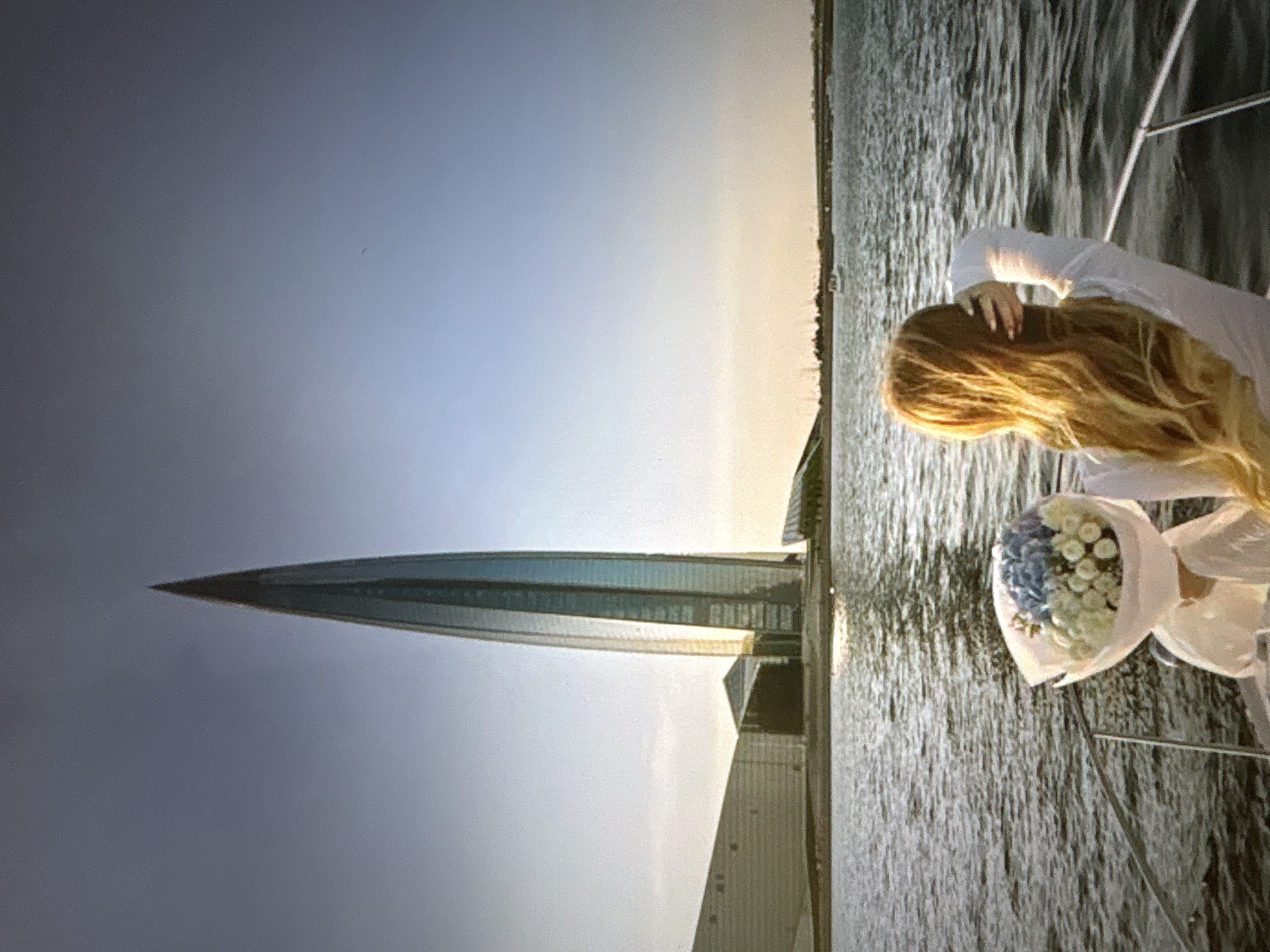In a wide shot, a woman with long, blonde hair is seen from behind, standing on the deck of a boat. She is wearing a white top and has her right hand raised, touching her hair. In the foreground, a bouquet of blue and white flowers is visible, wrapped in translucent paper. The boat is on a body of water, likely a bay or a river, with gentle ripples.
In the background, a modern, tall skyscraper with a pointed, glass facade dominates the skyline. Adjacent to the skyscraper are other contemporary buildings with angular designs. The architecture suggests an urban setting. The sky is a gradient of pale blue and orange, indicating either dawn or dusk. The scene appears to be in an urban waterfront area, and the architectural style is not typical of Pori, Finland, which is a smaller city known for its historical buildings and natural landscapes rather than such prominent modern skyscrapers. The prominent skyscraper is characteristic of a major metropolitan center.