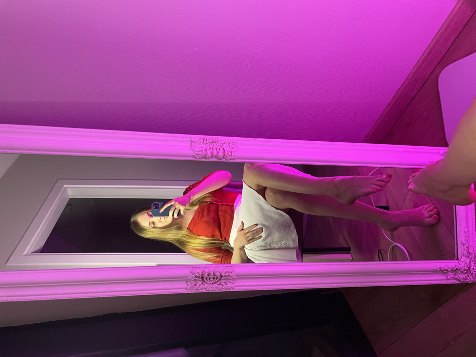 The image displays an indoor scene dominated by a large, ornate white-framed mirror. The entire room is illuminated with a strong, vibrant pink or magenta light, casting this color across the ceiling, the mirror frame, and parts of the floor.
Inside the mirror's reflection, a person is visible, seemingly taking a selfie. The person has long, straight blonde hair and is wearing a red ribbed top. A white piece of clothing, possibly a skirt or a towel, is draped over their lower body. They are holding a blue smartphone with their left hand, directing it towards themselves. Their bare legs are visible, one bent and the other extended, with bare feet showing red toenail polish. The person appears to be reclining or lying down, facing towards the right side of the frame as they capture their reflection.
The background reflected in the mirror includes a dark area, possibly a doorway or window opening, framed by white molding. A wooden floor is also visible in the reflection, along with a white cable resting on it.
Outside the mirror's frame, in the top portion of the image, the textured ceiling is brightly lit with the pink light. In the bottom right corner, a portion of a bare leg and foot, also with red toenail polish, is visible on a wooden floor, suggesting another person or perhaps the photographer's own foot is positioned outside the mirror's viewable area.
The overall impression is a casual, personal moment captured in a mirror selfie, characterized by the distinct and atmospheric pink lighting. No text is visible in the image.