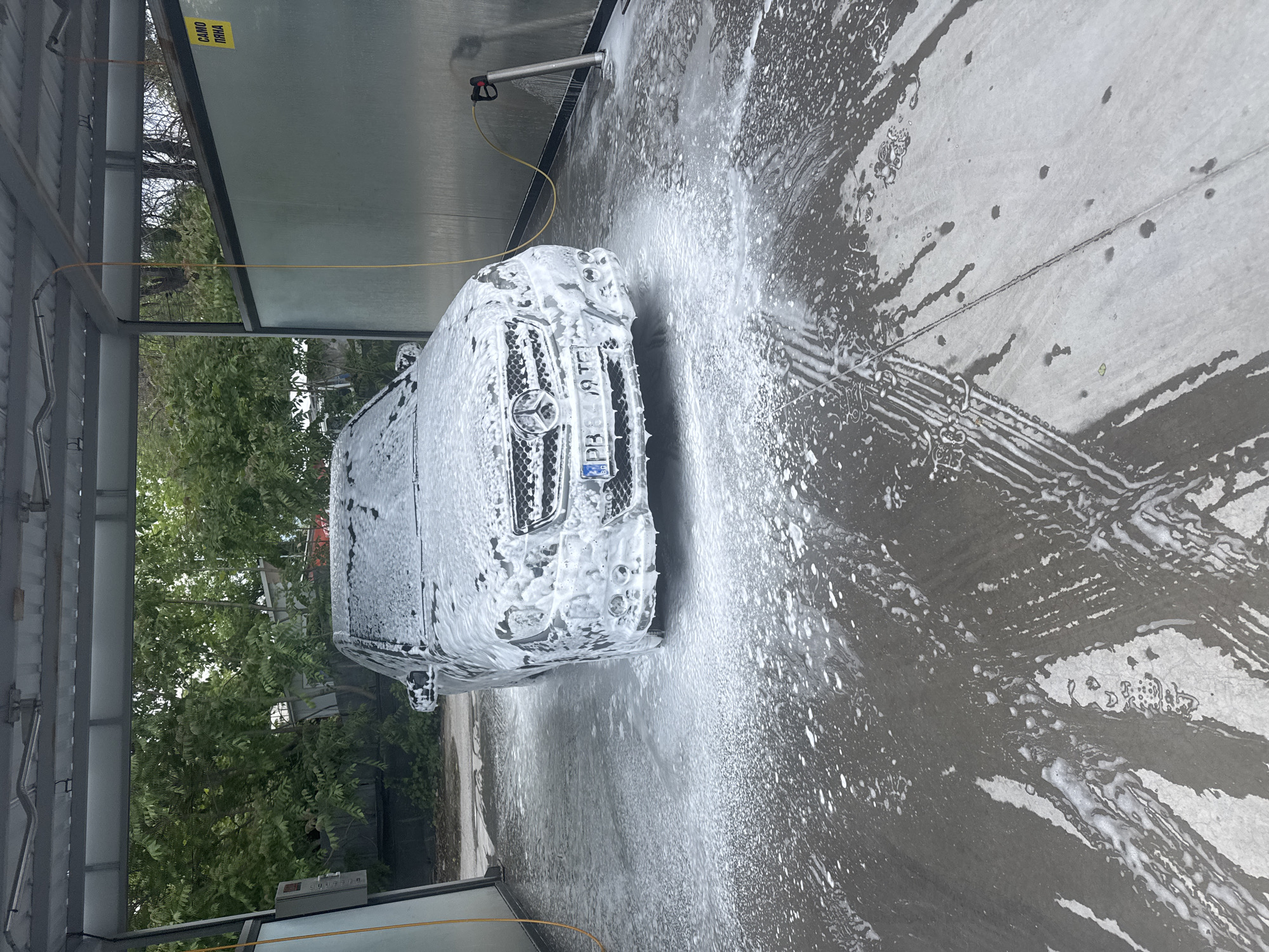 A car is being washed at a self-service car wash. The car is covered in white foam. A pressure washer wand is visible above the car, and water and foam are spraying onto the ground. The car has a Bulgarian license plate "PB 8419 TE". The car wash is located outdoors, with trees visible in the background. The location is likely Plovdiv, Bulgaria, based on the Bulgarian license plate and the request for the location.