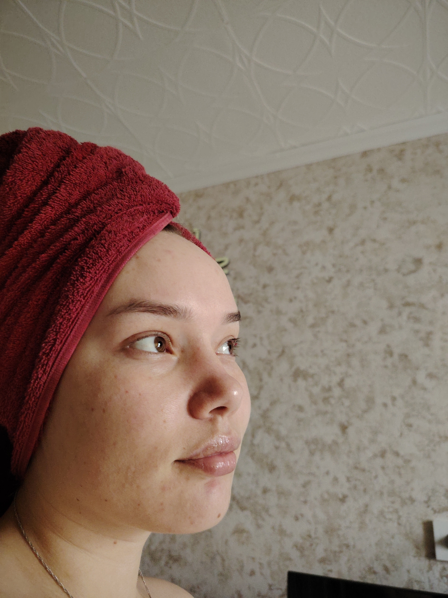 A woman with a red towel wrapped around her head is seen in a close-up, side-profile shot. She is facing right and looking upwards with light brown eyes. Her skin has a fair complexion with some freckles and minor blemishes visible. Her lips have a glossy appearance, possibly from lip gloss or balm. She appears to be indoors, with the background showing a textured wall and a ceiling with a circular pattern. A silver necklace is visible around her neck. Based on the available information, the location in the city of Марковский, Russia cannot be verified.