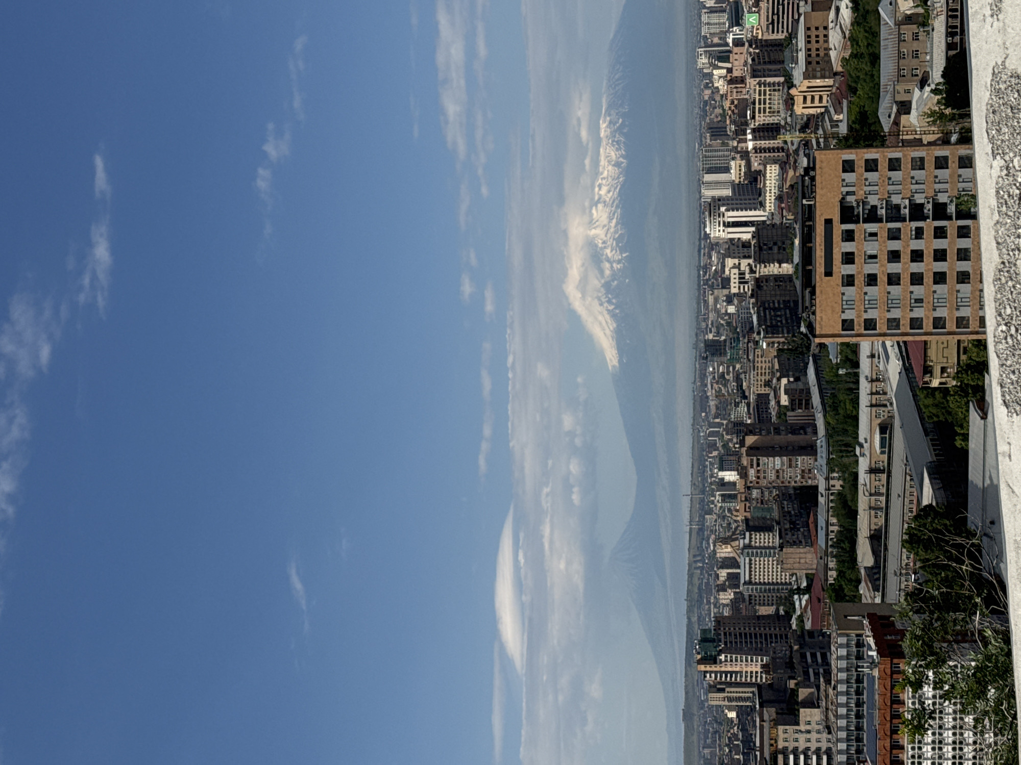 The image, taken from an elevated vantage point in Yerevan, Armenia, presents a sprawling urban landscape under a clear blue sky with scattered white, wispy clouds. The foreground shows the top edge of a building or balcony, hinting at the height from which the photo was captured.
The mid-ground is dominated by a dense array of city buildings, exhibiting various architectural styles and heights, with many featuring numerous windows. Some patches of green trees are visible between the structures, adding a touch of nature to the urban sprawl. The city extends far into the distance, appearing somewhat hazy.
Prominently featured in the background is a large, majestic snow-capped mountain, characteristic of Mount Ararat, partially veiled by atmospheric haze but still distinctly visible. The time of day appears to be bright daylight, possibly midday or early afternoon, given the strong illumination and clear sky. The weather is clear and sunny. No people or visible text are present in the image.