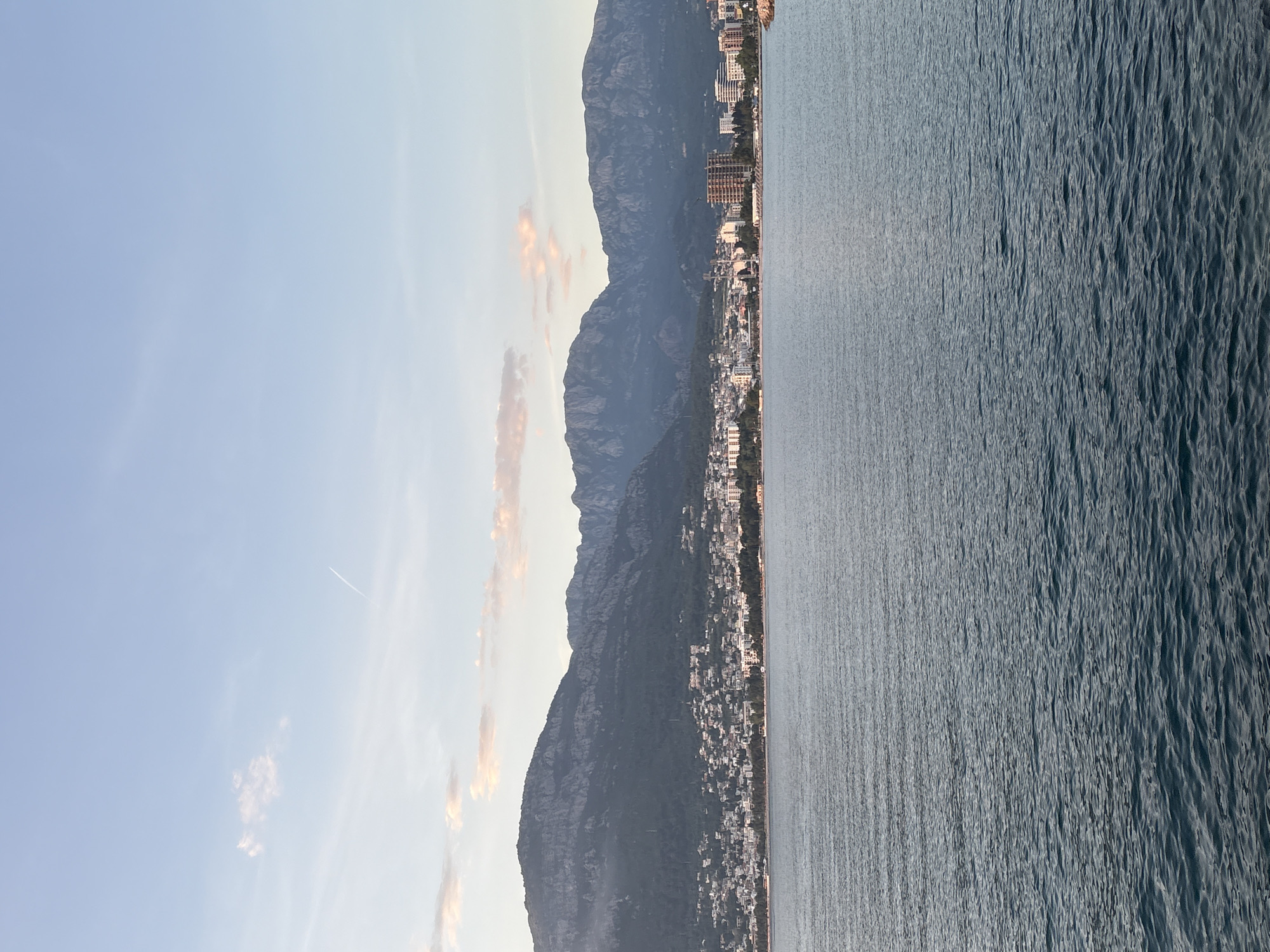 The image depicts a view of Bar, Montenegro, from the water, oriented with the coastline running vertically. A range of dark, mountainous terrain rises parallel to the city's coastal development. The water appears choppy, with distinct wave patterns. The sky is light blue, fading towards the horizon, with a few scattered clouds visible. A contrail is visible in the upper-left quadrant of the sky. Buildings of various sizes are densely packed along the coastline, transitioning from low-rise residential to taller structures as the city extends. The lighting suggests the photograph was taken during early morning or late afternoon, with soft, diffused light. There are no people immediately visible. The landscape suggests a Mediterranean coastal environment.