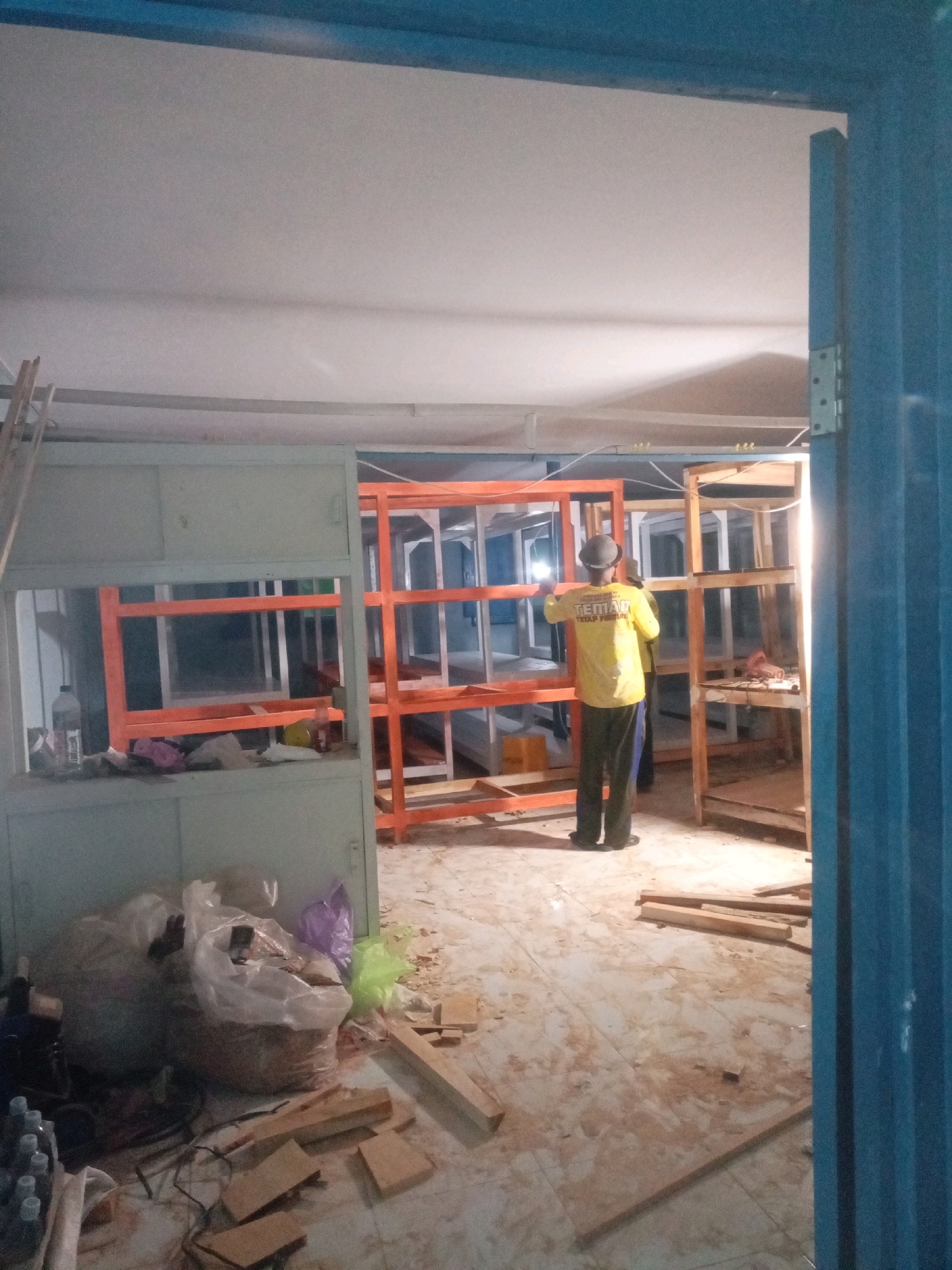 The image depicts an interior scene, likely a workshop or construction site, located in Sumbersari, Indonesia. A man wearing a yellow shirt with text and a hat is standing and working on an orange metal shelving unit. Another person is partially visible behind him, also appearing to be involved in the work. The environment is cluttered with construction materials, including lumber, plastic bags filled with debris, and various tools on the floor. The shelving units are unassembled or partially assembled. The lighting suggests it is either nighttime or an indoor space with artificial illumination. The floor is tiled and appears dusty and unkempt, consistent with an active work area. A blue metal door frame is visible on the right side of the image, suggesting an entrance or exit.