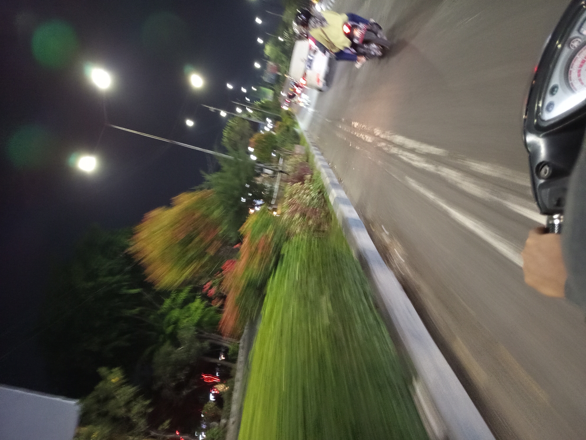 This is a first-person perspective, night-time photograph taken from a moving motorcycle on a wet asphalt road in Buduran, Indonesia. The road is illuminated by streetlights and the headlights of other vehicles. In the foreground, the hand of the rider is visible, gripping the handlebars of the motorcycle, which features a digital dashboard. Further ahead on the right side of the road, another motorcycle with two riders, one wearing a yellow jacket and a helmet, is also moving. On the left side of the road, a green grassy area with some flowering bushes and trees is visible, along with streetlights illuminating the scene. The motion blur indicates the photograph was taken while the motorcycle was in motion, suggesting travel through an urban or suburban area at night.