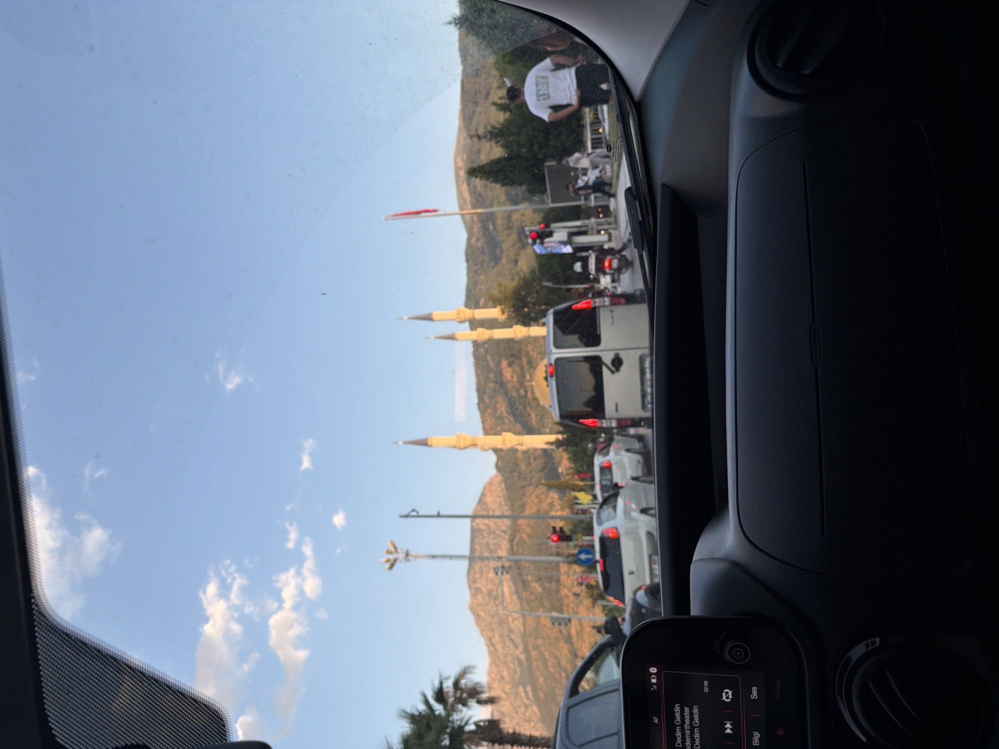 The image was taken from inside a car, looking out through the windshield. The foreground shows the dashboard of the car, including a digital display screen showing song information. The middle ground depicts a street scene with several cars, some in motion and some stopped in traffic. A person in a white t-shirt and black shorts is visible standing on the right side of the road, facing away from the camera. In the background, there are two mosque minarets and a dome, with a mountain range behind them. The sky is blue with scattered clouds. The location is identified as Yunus Emre, Turkey. The overall atmosphere suggests a busy urban environment during daylight hours, likely late afternoon or early evening due to the lighting.