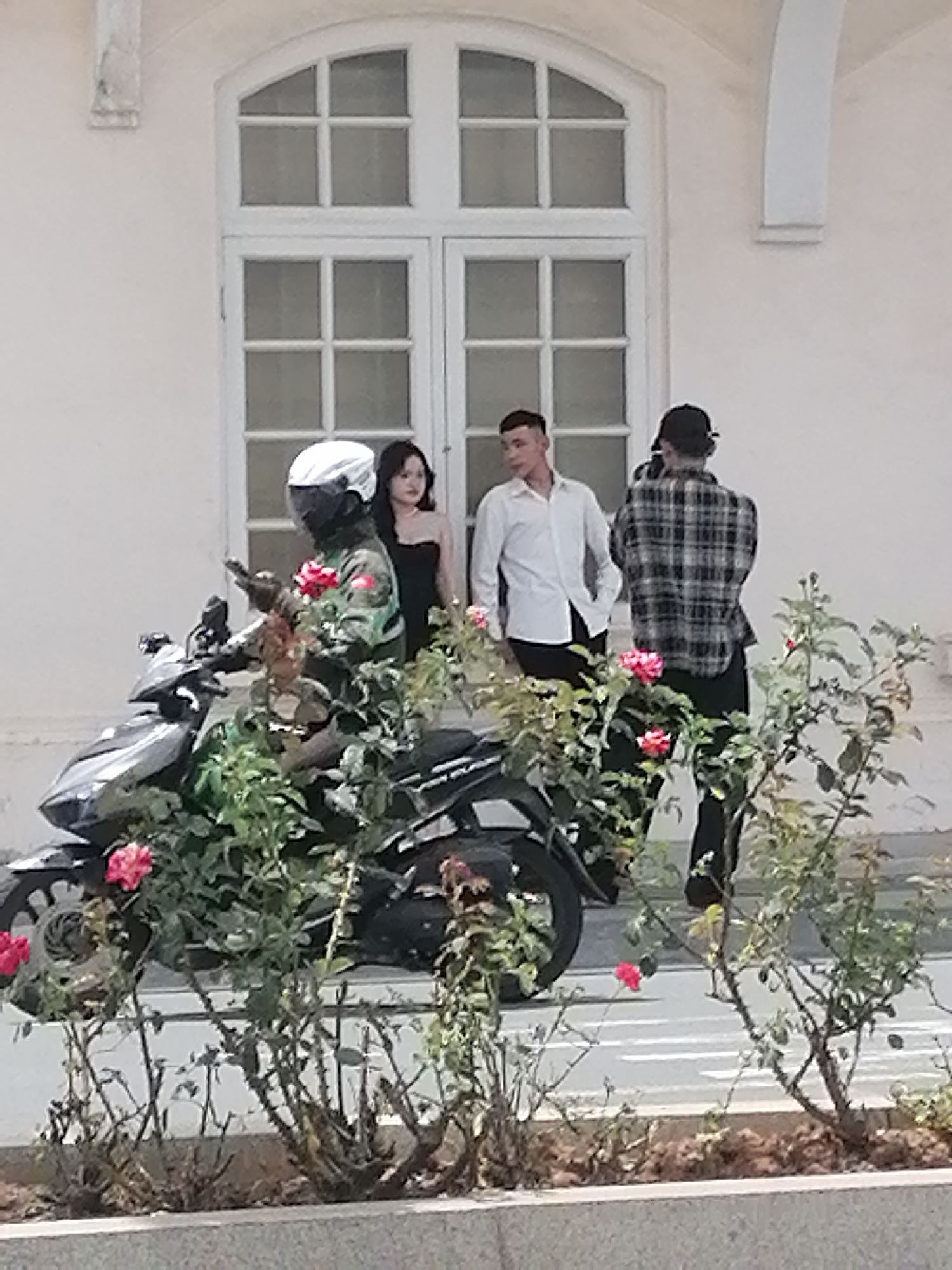 The image depicts a scene in front of a white building with a large window. On the left, a person wearing a camouflage jacket and a white helmet is seated on a black scooter. The person appears to be holding a phone. Next to them, a woman in a black strapless dress stands looking away. To the right of the woman, a man in a white collared shirt and black pants is standing with his hands in his pockets. Further to the right, another person wearing a plaid shirt and a black baseball cap is visible from the back. Rose bushes with pink flowers are in the foreground, obscuring the lower part of the scooter and the building's base. The background shows a street with white lines. The architecture of the building, particularly the window and the decorative elements, suggests it could be an older, possibly colonial-style building in Da Lat, Vietnam, known for its French colonial architecture and many floral gardens.