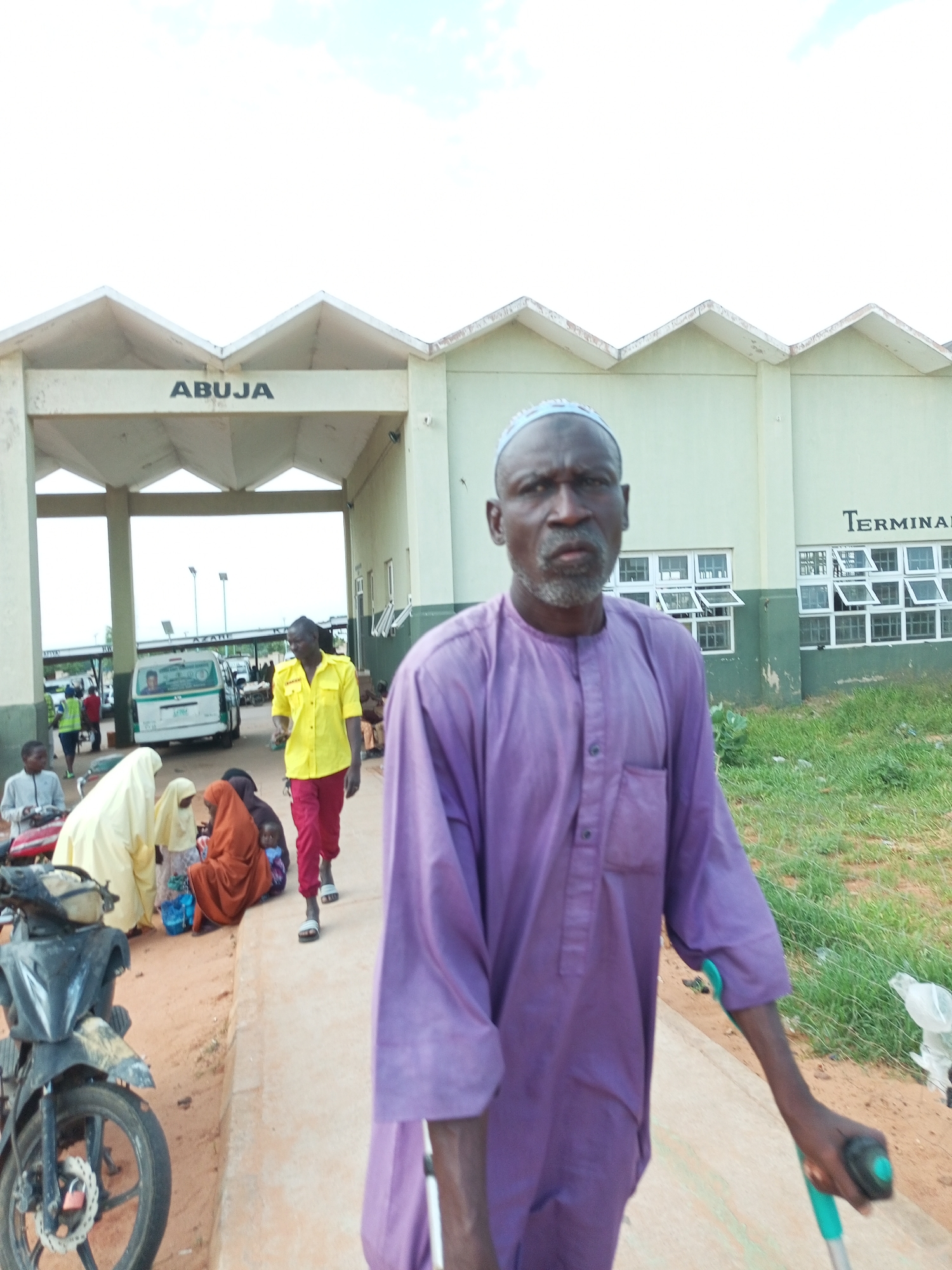 A man with a grey beard and wearing a purple robe and a white skullcap is the central figure in the foreground of this image, walking with the aid of a blue and green crutch. He is facing the camera with a serious expression. Behind him, in the mid-ground, a building with "ABUJA" written above its entrance is visible. To the left of the building, there's a beige-colored structure with the word "TERMINAL" visible on it. Several people are gathered in front of the building. A man in a bright yellow shirt and red trousers walks towards the right side of the frame. Others, including women in headscarves and children, are seated or standing near a motorcycle. The ground is a mix of dirt and a paved walkway. The environment appears to be an outdoor public space, possibly a transportation terminal. The sky above is mostly white with some clouds. The image likely depicts the Abuja transport terminal.