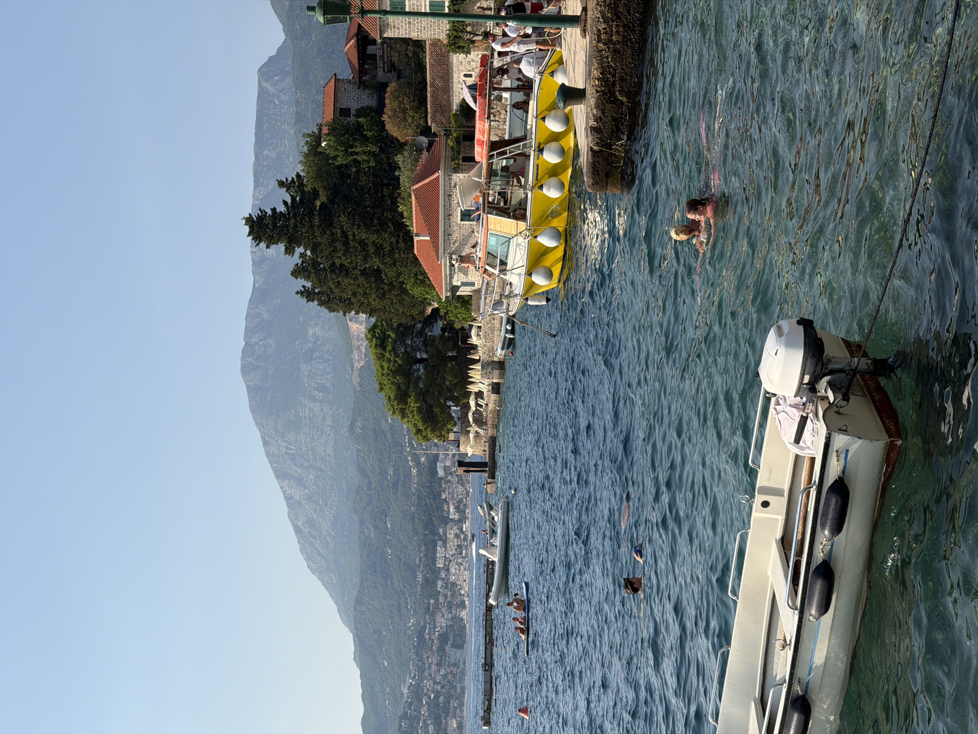 The image captures a sunlit coastal scene in Zelenika, Montenegro, with a clear blue sky and tranquil, turquoise water. In the foreground, a white motorboat with a white outboard motor is moored, its hull showing a red waterline and black fenders. Further into the water, two people are swimming; one appears to be a man wearing a hat, holding a ball, while a second person's head is visible near him. In the mid-ground, a group of people are on a small inflatable raft. To the right, a yellow passenger boat with a canopy is docked at a stone pier, with several people visible on and around it. Behind the pier, stone buildings with red-tiled roofs are visible, nestled amongst lush green trees. The background features a densely populated hillside with buildings cascading down towards the water, and a large, imposing mountain range under the clear sky. The overall atmosphere is one of leisure and summer activity by the Adriatic Sea.
