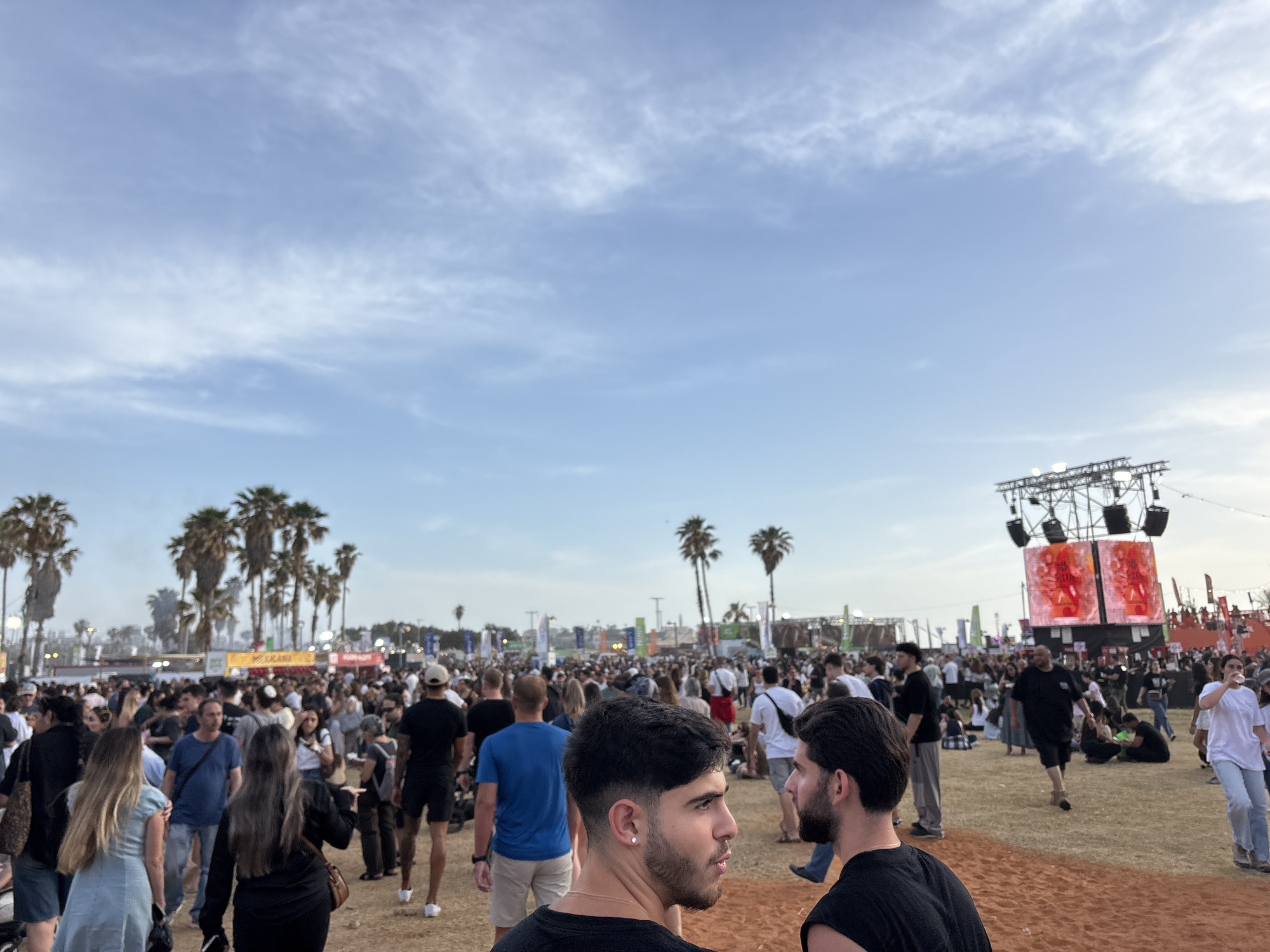 The image depicts a crowd of people at a large outdoor event. The location appears to be a park or open space, with palm trees scattered throughout the area and a sandy ground surface. The crowd is dense and varied, with individuals of different ages and ethnicities dressed in casual clothing. In the background, there is a stage structure with large screens displaying images or videos. The sky is light blue with some clouds. Two young men are in the foreground, facing away from the camera, partially obstructing the view. Based on the presence of palm trees and the general atmosphere, the location could be in Tel Aviv, Israel.