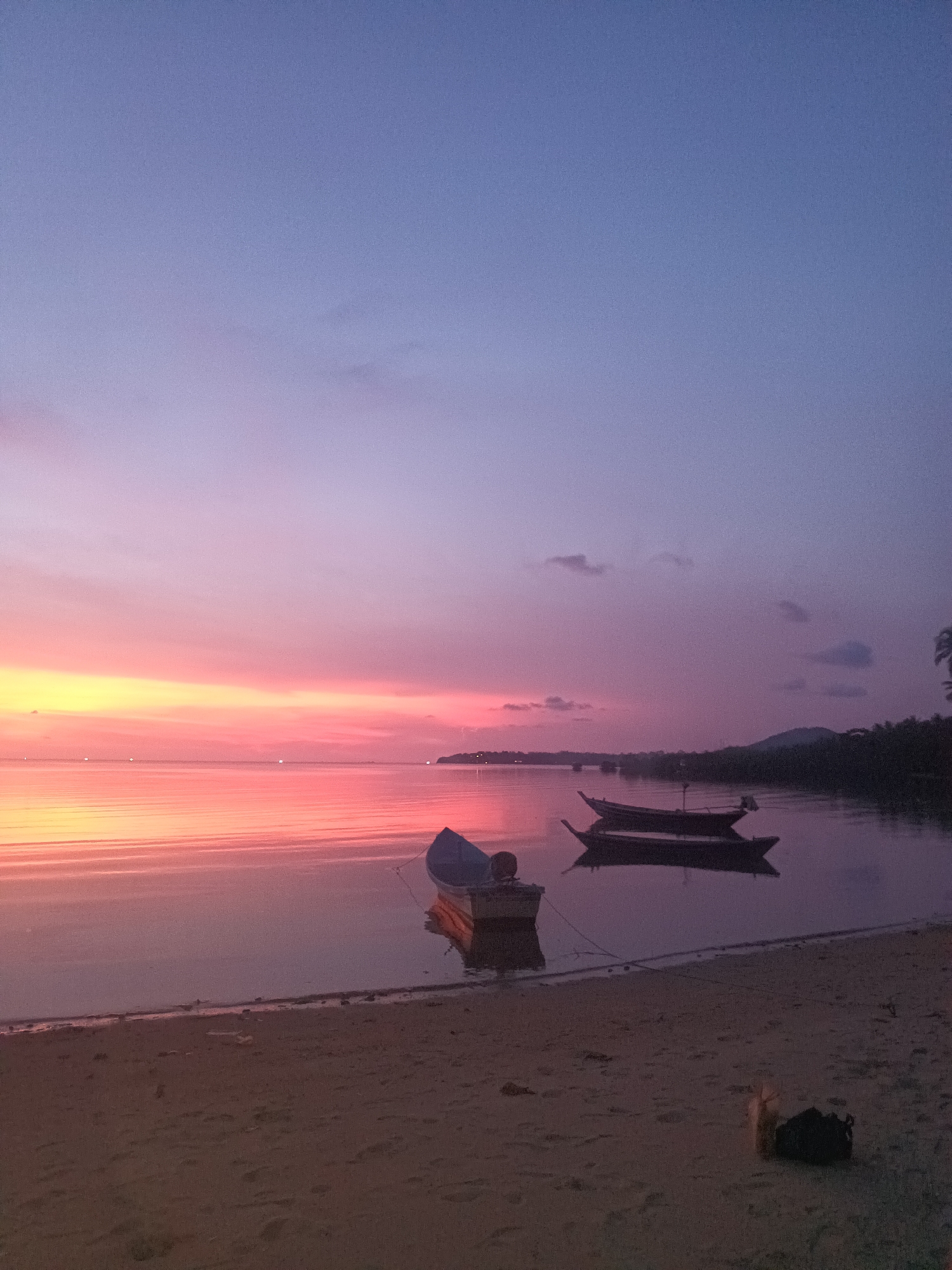 The image depicts a serene sunset over a body of water, likely the sea, in Baan Wok Tum, Thailand. The sky transitions from a light blue at the top to a vibrant mix of pinks, oranges, and yellows near the horizon, which reflects on the calm water surface. Three boats are visible: one close to the shore, seemingly beached, and two others further out, floating on the water. The shoreline is sandy, with some debris and vegetation visible. The distant shoreline shows signs of habitation and possibly a mountain range.