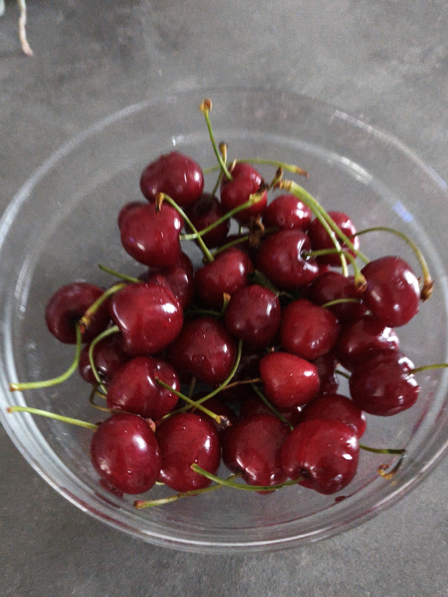 A close-up, overhead shot displays a clear glass bowl filled with dark red cherries. The cherries have visible stems, and some appear to be slightly wet. The bowl is placed on a textured, grey surface. The scene is lit with soft, diffused light, creating subtle reflections on the cherries and the bowl. There are no people or animals present in the image. The background is out of focus, suggesting a shallow depth of field. The image does not contain any information that would allow for the identification of its location in the city of Brodi, Latvia.