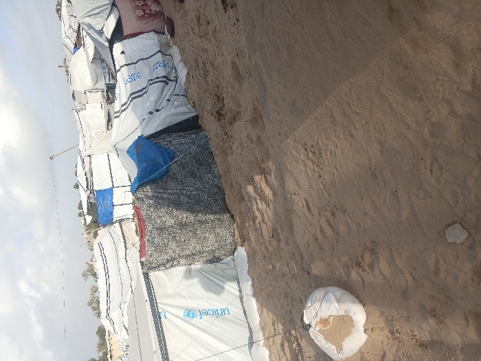 The image depicts a crowded, makeshift displacement camp situated on sandy terrain in Khan Yunis, Palestinian Territory, under a partially cloudy sky. The foreground shows light brown sand with footprints and a small, light-colored stone. A white sandbag, partially open and filled with sand, lies in the lower-left, tethered by a dark rope extending upwards, likely anchoring a shelter. The midground and background are dominated by numerous closely packed shelters constructed from white and blue tarpaulins, some bearing the "Unicef" logo. Other shelter materials include dark grey patterned fabric and patterned cloth, such as a red and white floral design on the right. The shelters exhibit varied configurations and appear to be temporary structures. No individuals are visible. Sparse vegetation is present in the distant background beyond the dense array of tents.