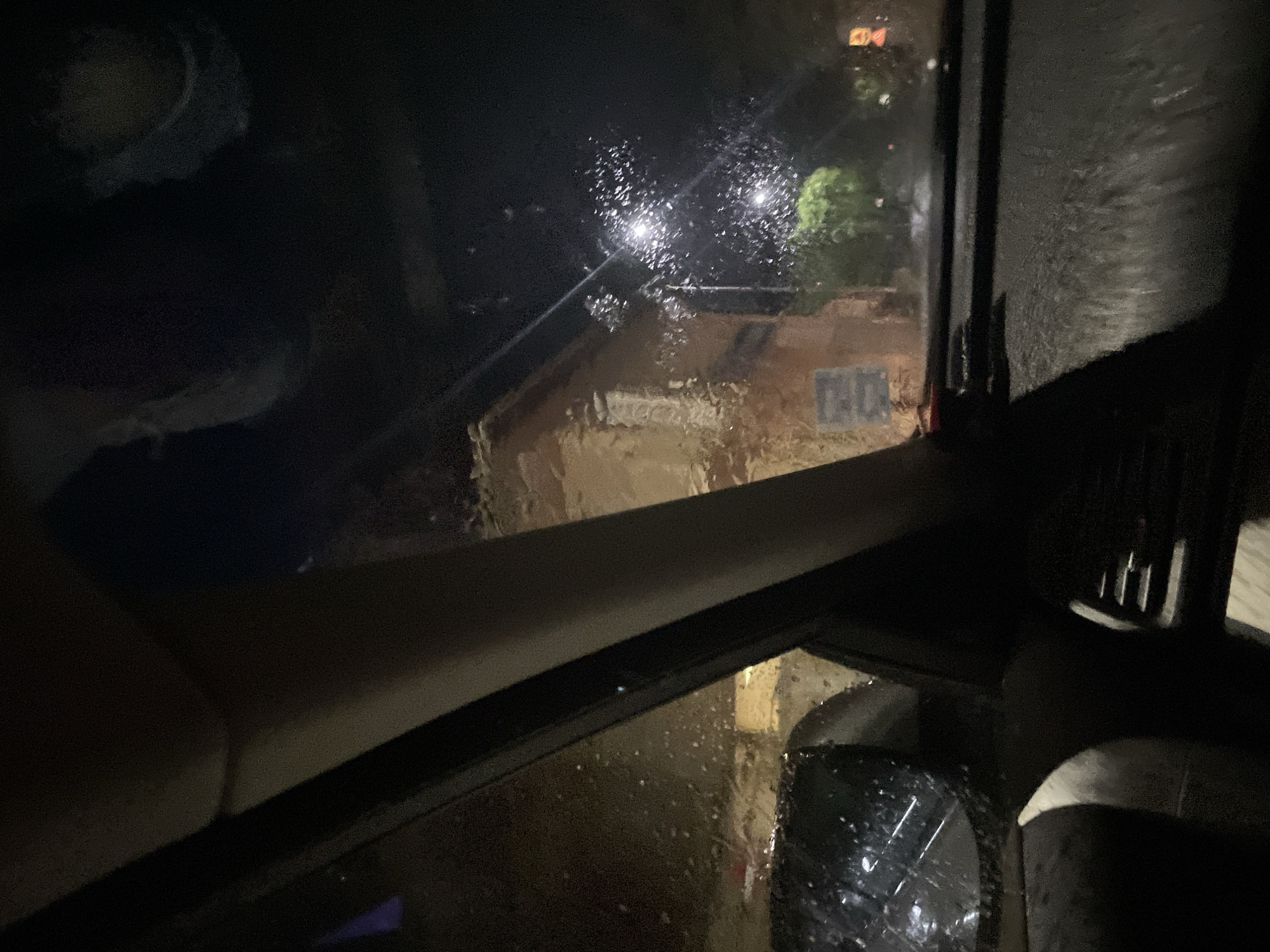 The image shows a view from inside a vehicle during a rainy night in what is believed to be Zlatograd, Bulgaria. Raindrops cover the car window, obscuring the view of a building across the street, illuminated by streetlights. The building appears to be made of brick and has some sort of signage on it. Parts of the car's interior are visible, including the dashboard, side mirror, and a partially visible passenger.