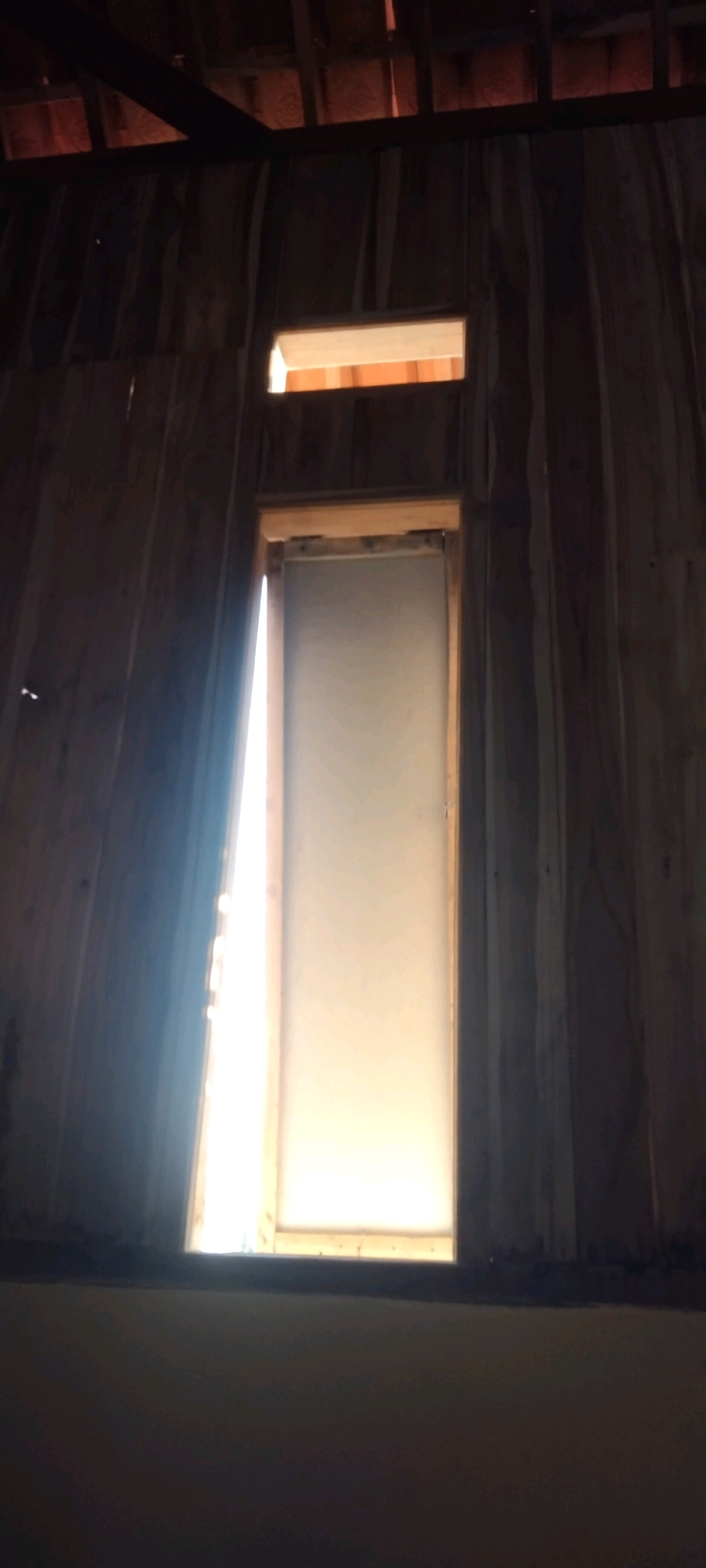 The image depicts the interior of a dimly lit structure, likely under construction or in a state of disrepair, with visible wooden beams and paneling. A large, rectangular opening, resembling a window or doorway, dominates the center of the frame. This opening is covered by a translucent screen, through which bright light is emanating, suggesting it is facing an outdoor light source. The light spills dramatically from the left side of the opening, casting a bright glow and creating a strong contrast with the shadowed interior. Above this main opening, a smaller rectangular opening is visible, also letting in light. The wooden walls are roughly finished and appear to curve inwards towards the top, possibly indicating a circular or semi-circular roof structure supported by beams. The floor area at the bottom of the image is dark and out of focus. The overall impression is of an unfinished interior space illuminated by external light filtering through an opening. No people, animals, or specific identifiable objects are present, beyond the structural elements of the building. The scene does not provide any specific clues to its location in Unknown, Indonesia.