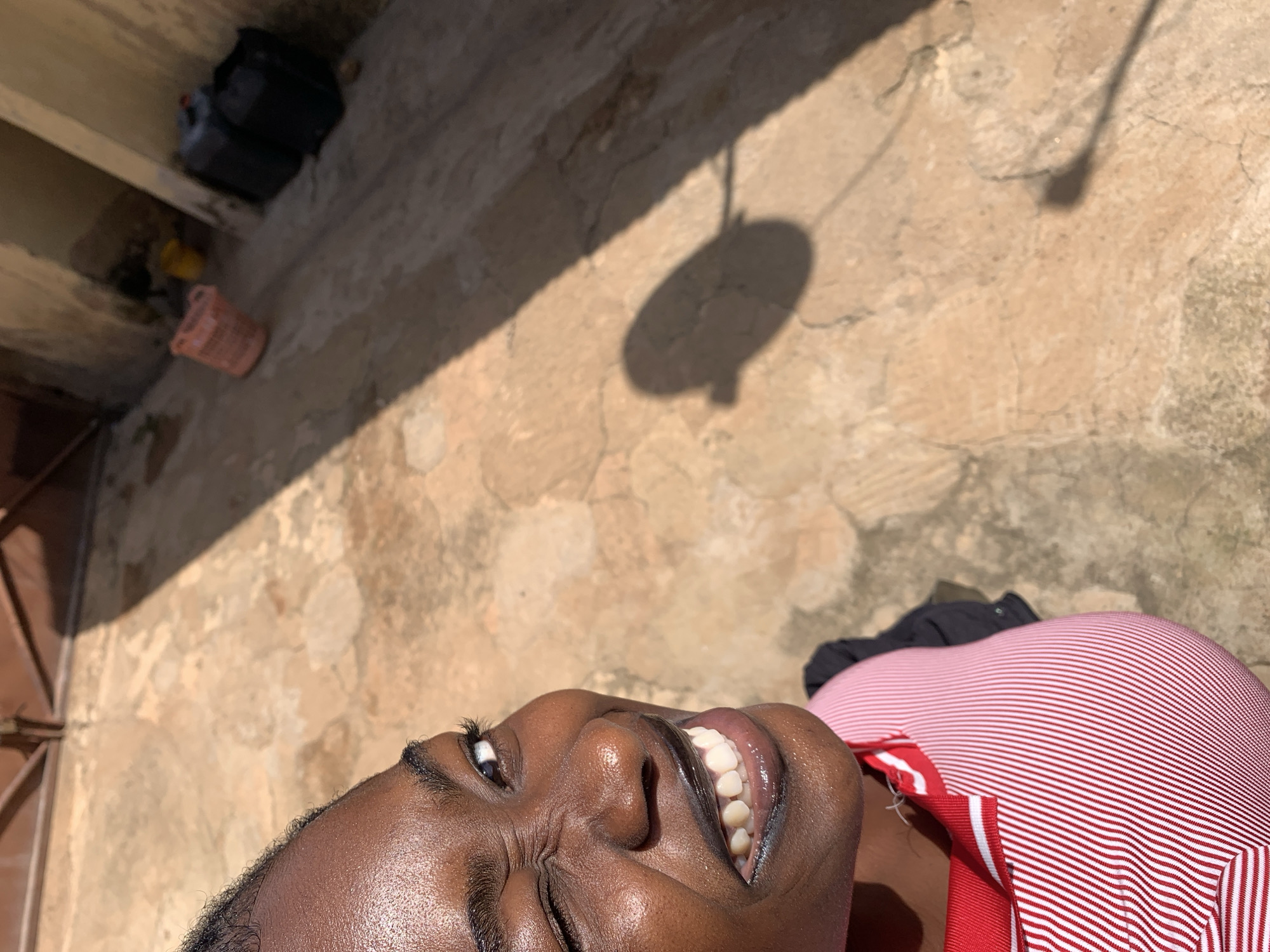 The image captures a close-up of a Black woman with dark skin, winking and smiling broadly, with her teeth visible. She is wearing a red and white striped shirt. The background appears to be an outdoor, possibly urban, setting with a concrete ground, a wall, and shadows. In the background, to the right, are two dark plastic containers and a pink basket. The lighting suggests a sunny day. The scene is consistent with the environment found in Rafin Par, Nigeria, given its typical urban and semi-urban landscapes.