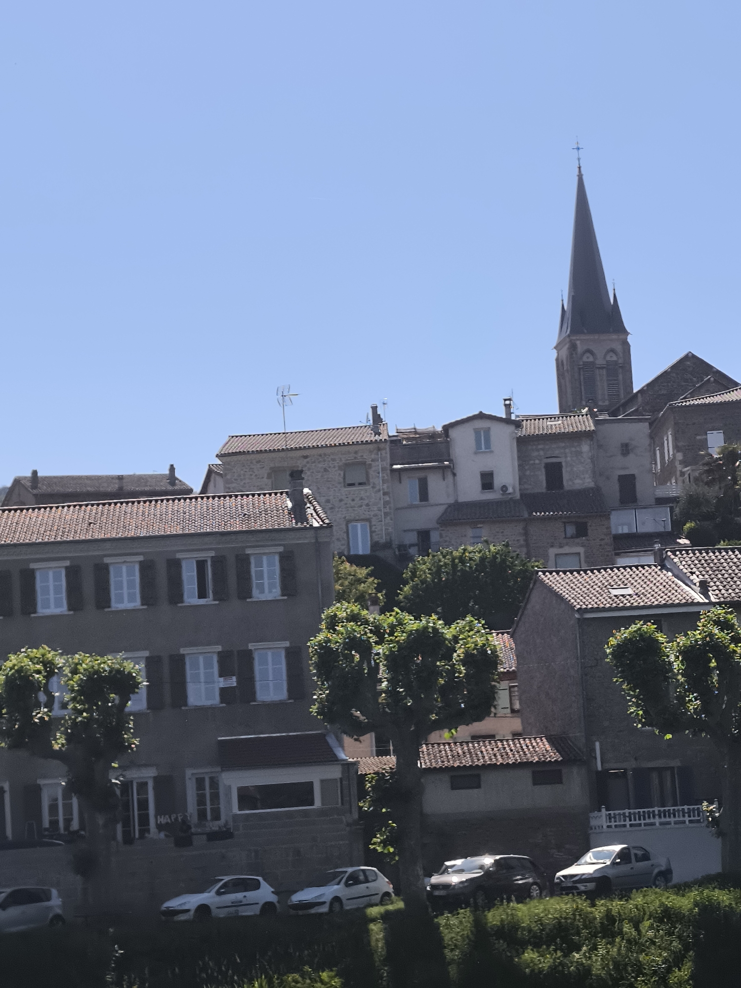 The image captures a section of Saint-Pierre-de-Bœuf, France. A low-angle shot reveals a variety of buildings, including residences and a prominent church steeple against a clear blue sky. The architecture is mixed, with some structures featuring red-tiled roofs and light-colored stone facades. Windows with dark shutters are visible. The steeple of the church is a dark color. Trees with rounded canopies are planted along the street, partially obscuring the lower portions of the buildings. Parked cars line the street in front of the buildings. Vegetation is visible in the foreground. The buildings ascend a slight hill. The word "HAPPY" can be seen painted on a wall.