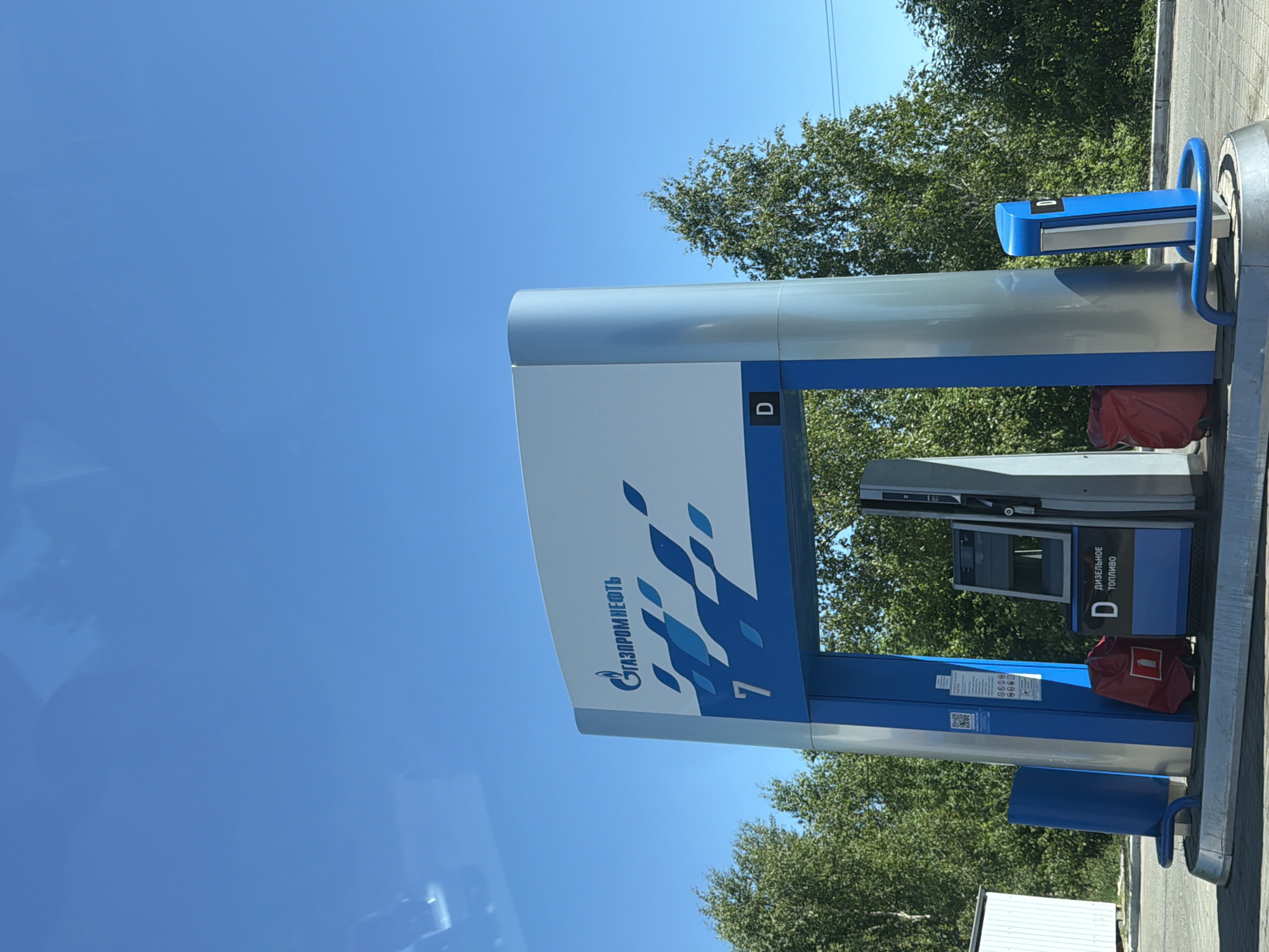 A clear, bright outdoor daytime photograph captures a modern Gazpromneft gas station fueling island. The central structure features a large white sign displaying the "ГАЗПРОМНЕФТЬ" (Gazpromneft) logo, blue geometric patterns, the number "7", and a "D" indicating diesel fuel, all supported by prominent blue and silver pillars. A fuel pump, featuring a digital display and indicating "D" and "ДИЗЕЛЬНОЕ ТОПЛИВО" (Diesel Fuel), is positioned underneath the canopy. Two red fabric bags or covers rest on the paved ground next to the pump, one on each side. Blue metal protective barriers are installed around the base of the fueling island. To the far left, a small white corrugated metal shed is partially visible. The background consists of dense green deciduous trees under a vast, clear blue sky. No individuals are present in the scene. The location is in the city of Unknown, Unknown.