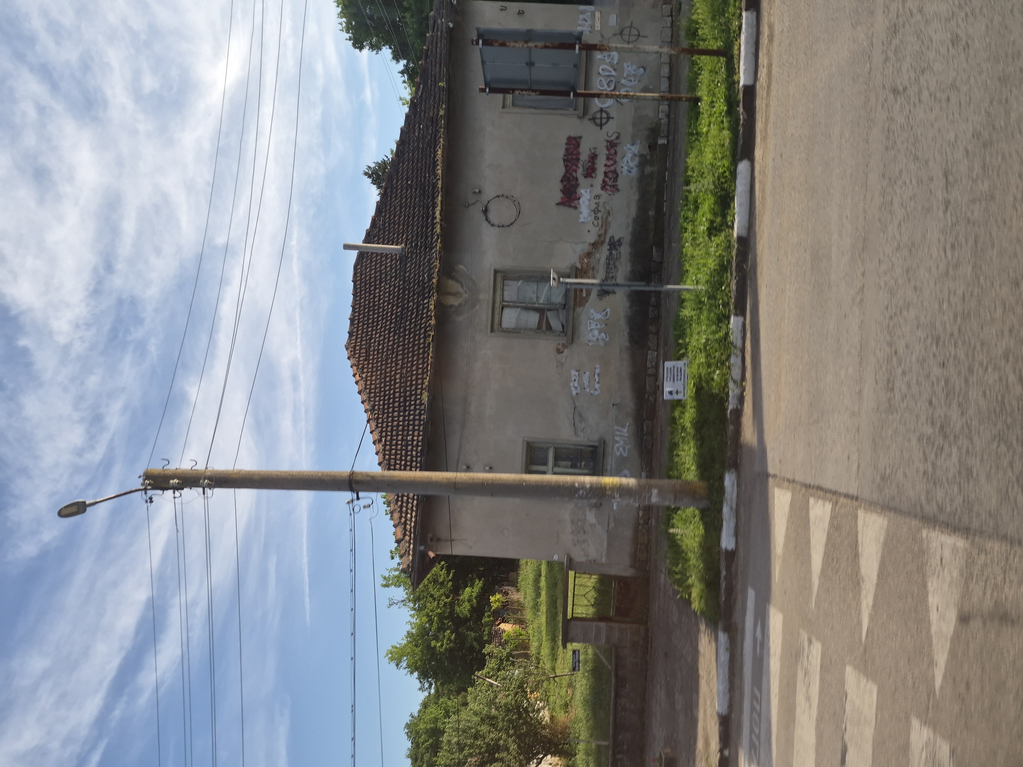 The image shows a view of a street in Balgarene, Bulgaria. On the left, a concrete utility pole with a street light stands prominently, with power lines extending across the frame. To the right of the pole is a one-story, weathered house with a red tile roof and visible signs of graffiti on its facade. The house has two windows on the visible side, one of which appears damaged. A crosswalk is visible in the foreground, indicating a pedestrian crossing area on the asphalt road. Behind the house, lush green trees and foliage form a backdrop, and a metal gate with a stone pillar is visible on the left side of the property. The sky is a clear blue with scattered white clouds.