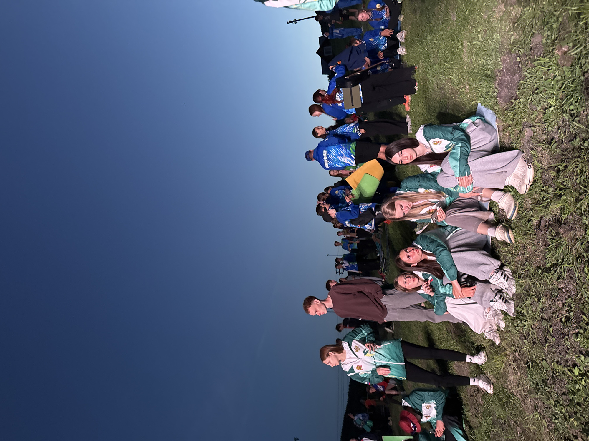 A group of people, including adults and teenagers, are gathered outdoors on a grassy area. Some are sitting or lying down, while others stand. Most appear to be looking in the same direction towards the left side of the image, possibly at something beyond the frame. The location appears to be Khvalynsk, Russia, as some people are wearing clothing with "Khvalynsk" printed on it. The sky is a deep blue, suggesting it is dusk or evening. There are power lines in the far background.