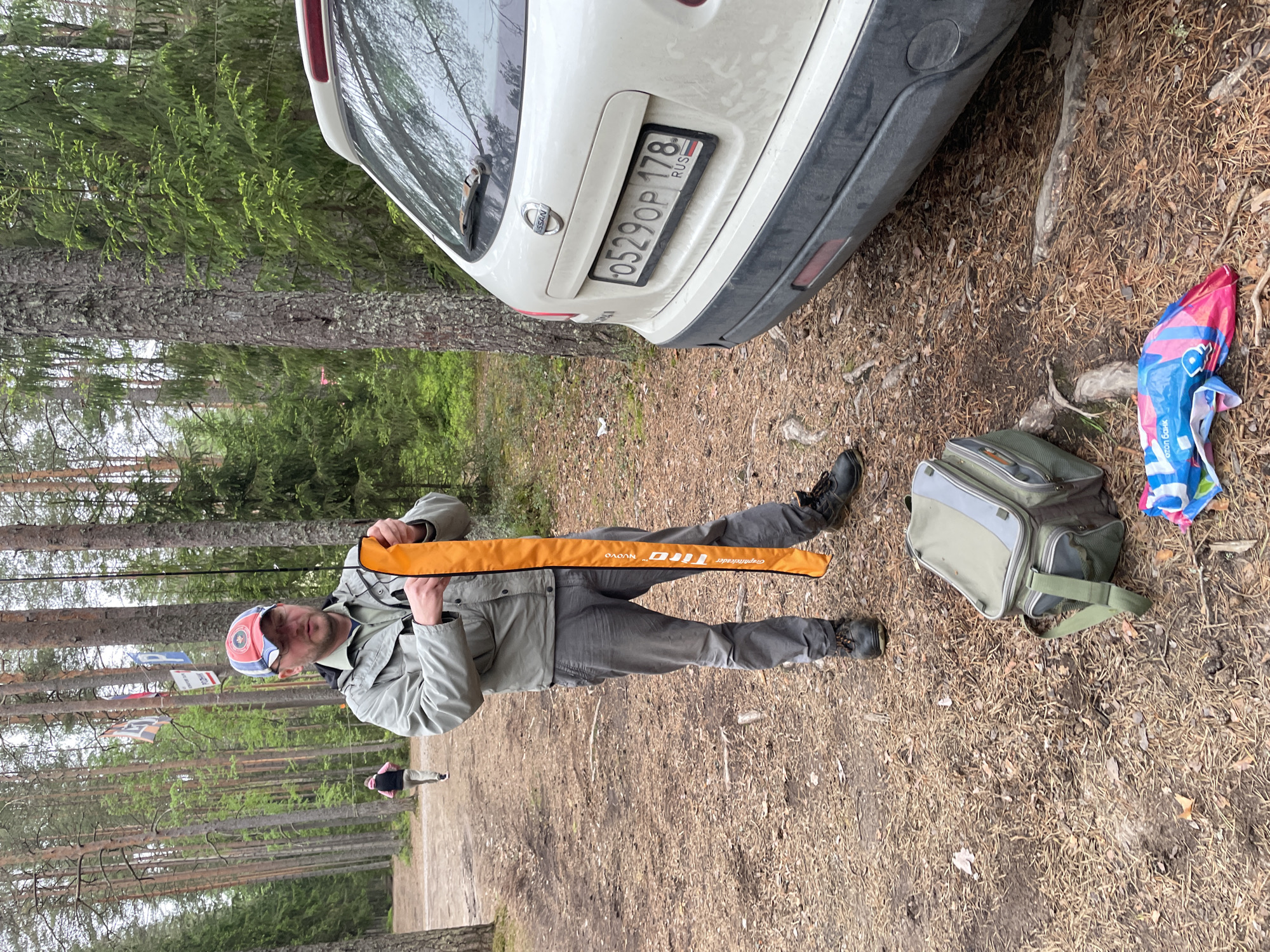 A man in gray attire stands in a forest area, holding a long orange case. He wears a multi-colored baseball cap and dark shoes. To his right, there's a light green bag and a blue and pink plastic bag. Behind him, a white car, partially visible, displays a Russian license plate "O529OP 178 RUS". The background consists of tall trees, suggesting a woodland environment. In the distance, another person is visible on a pathway. The scene appears to be in an area with pine needles and foliage on the ground.