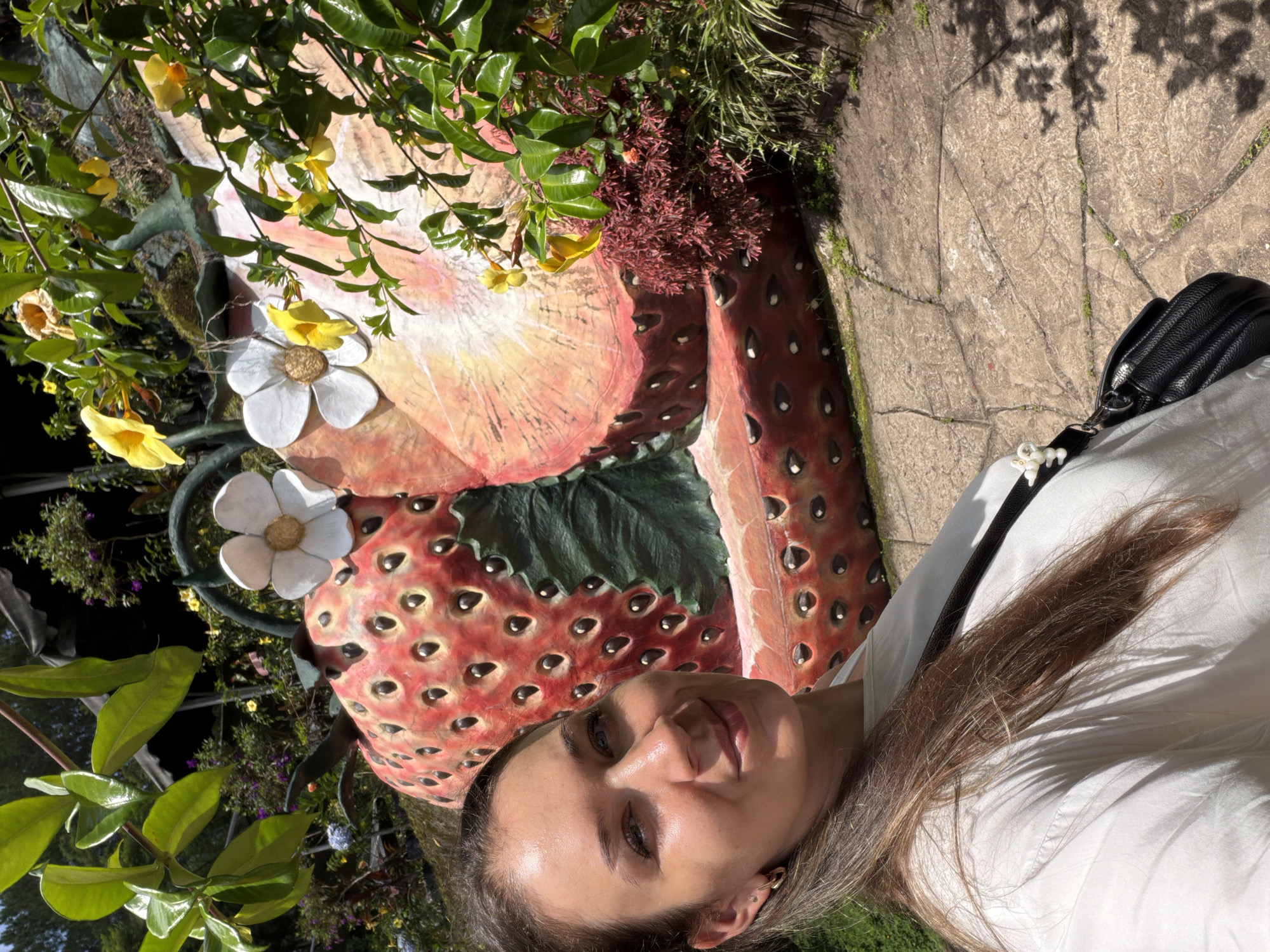 A young woman, with long brown hair, smiles at the camera in a selfie taken outdoors. She wears a white shirt and a black purse with white decorations hangs over her shoulder. Behind her, a giant sculpture of a sliced strawberry is adorned with white and yellow flowers and lush green foliage. The sculpture is situated on a paved pathway in what appears to be a garden or park, possibly in Da Lat, Vietnam, a city known for its strawberry farms and botanical gardens. The lighting suggests it is a sunny day.