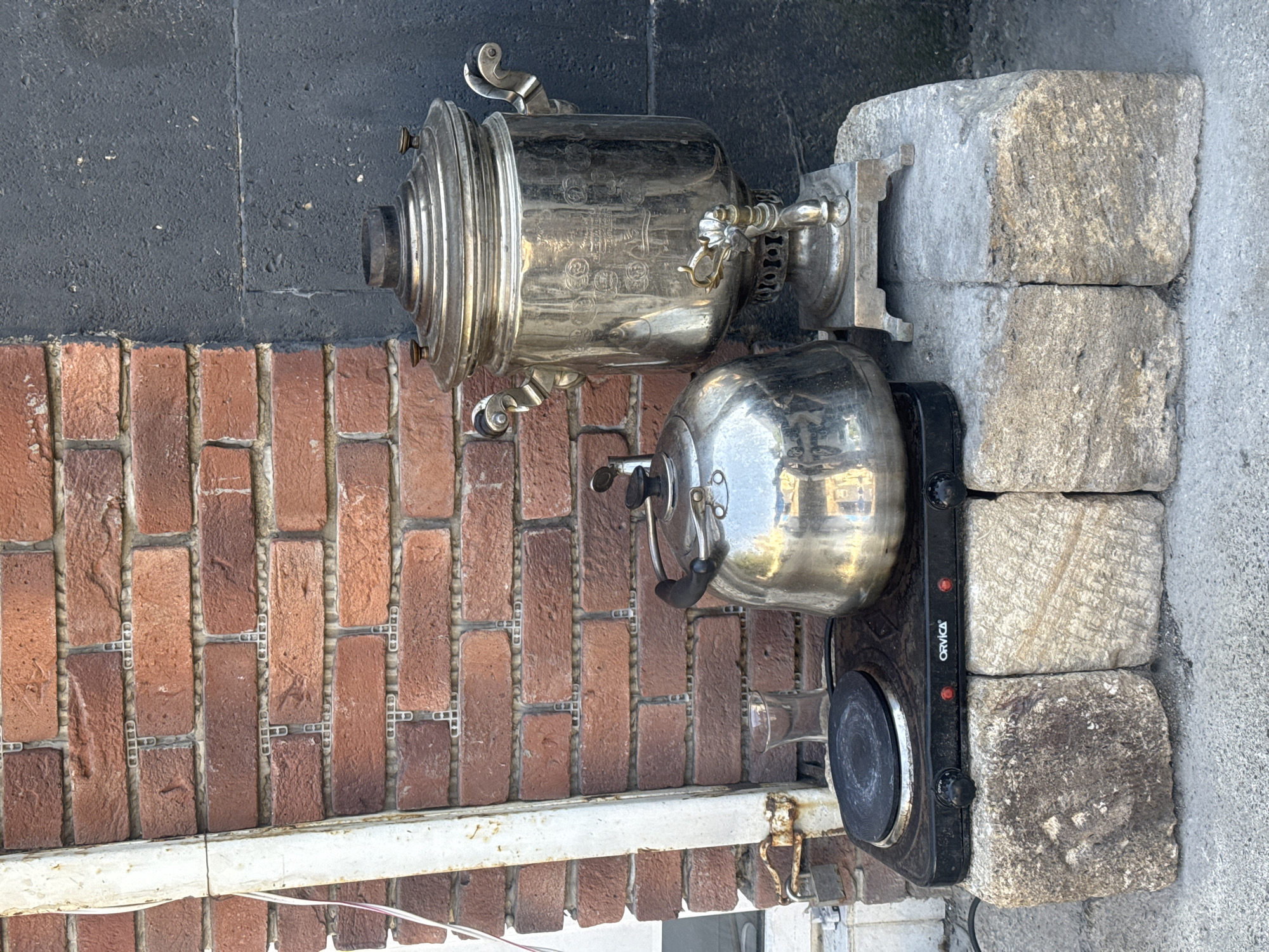 An outdoor scene in Baku, Azerbaijan, features a collection of items staged on rough-hewn stone blocks against a weathered brick wall and a dark grey textured wall. On the left, a small, black electric hot plate, branded "ORVICAL," sits on two of the stone blocks. A shiny metal kettle is positioned on the hot plate. To the right of the kettle, a clear glass is visible. Further to the right, a large, ornate, silver-colored samovar stands on its own stone block. The samovar has intricate detailing and a spigot. A white pipe with wires runs vertically along the left side of the brick wall. The ground is concrete. There are no people visible in the image.