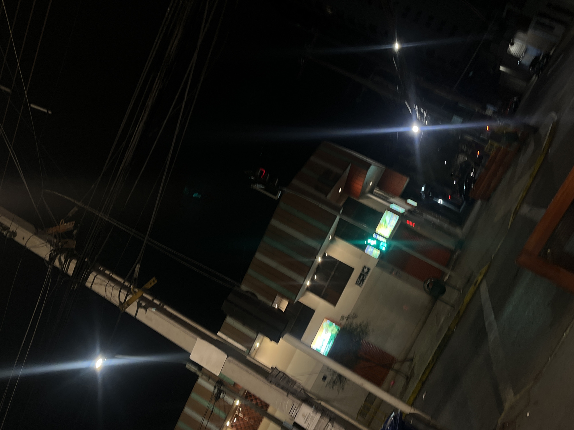 The image depicts a night scene on a city street in Iquique, Chile, likely near the Plaza Prat due to the presence of a prominent traffic light and buildings with awnings. A tall utility pole with numerous wires dominates the left side of the frame, with a street light casting a strong glare upwards and to the left. In the mid-ground, a building with an orange and green striped awning is visible, featuring illuminated signs and windows. Another building to the right appears to be part of the same complex. A traffic light, with its red light illuminated, is positioned on the right side of the street. The street is paved, with yellow lines marking lanes and the edge of the sidewalk. The overall scene is dark, with the artificial lights of the city providing the illumination. No people are clearly visible in the image.